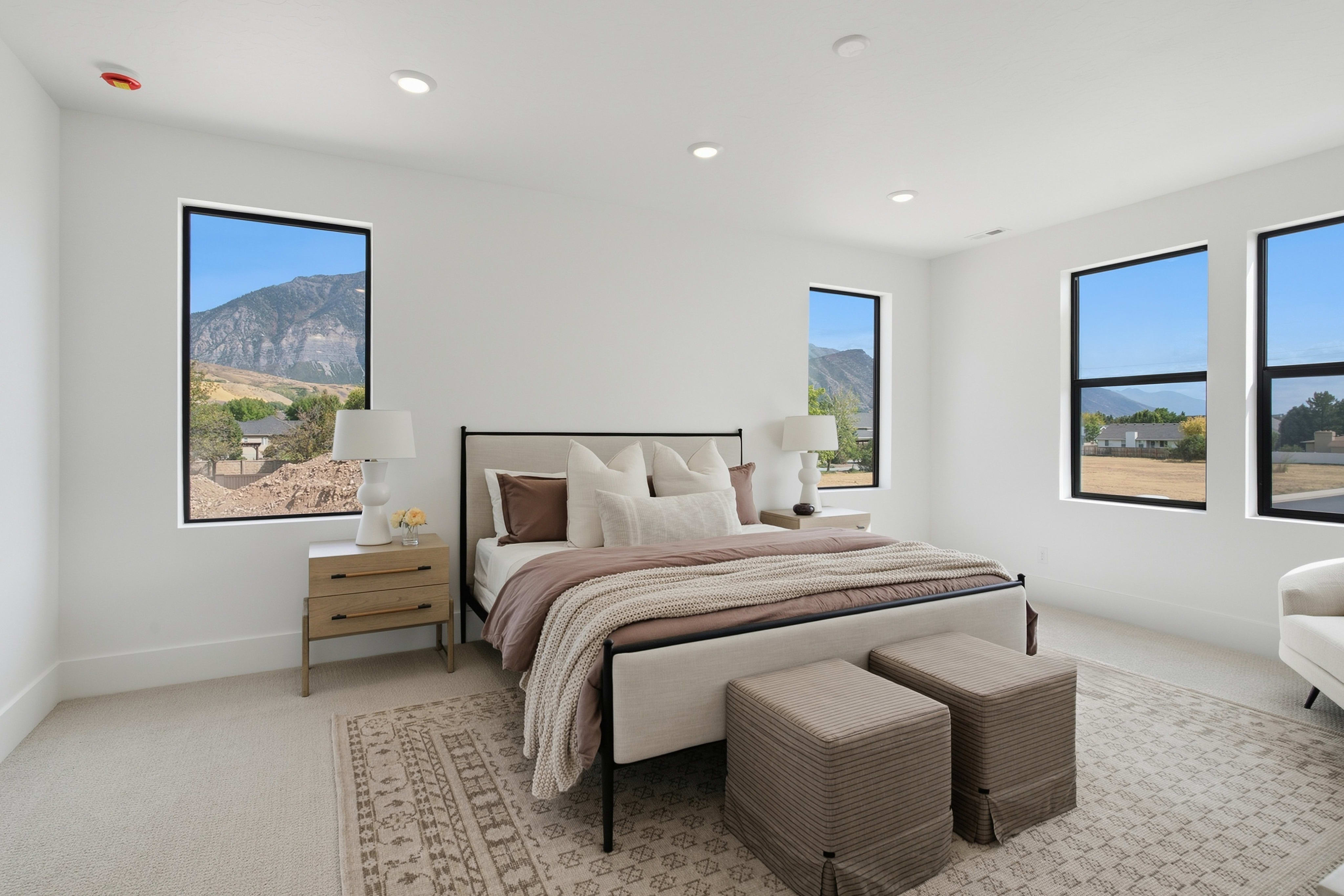 A modern bedroom features a bed with neutral linens and two ottomans in the foreground, set against white walls and windows overlooking a mountainous landscape in the background.
