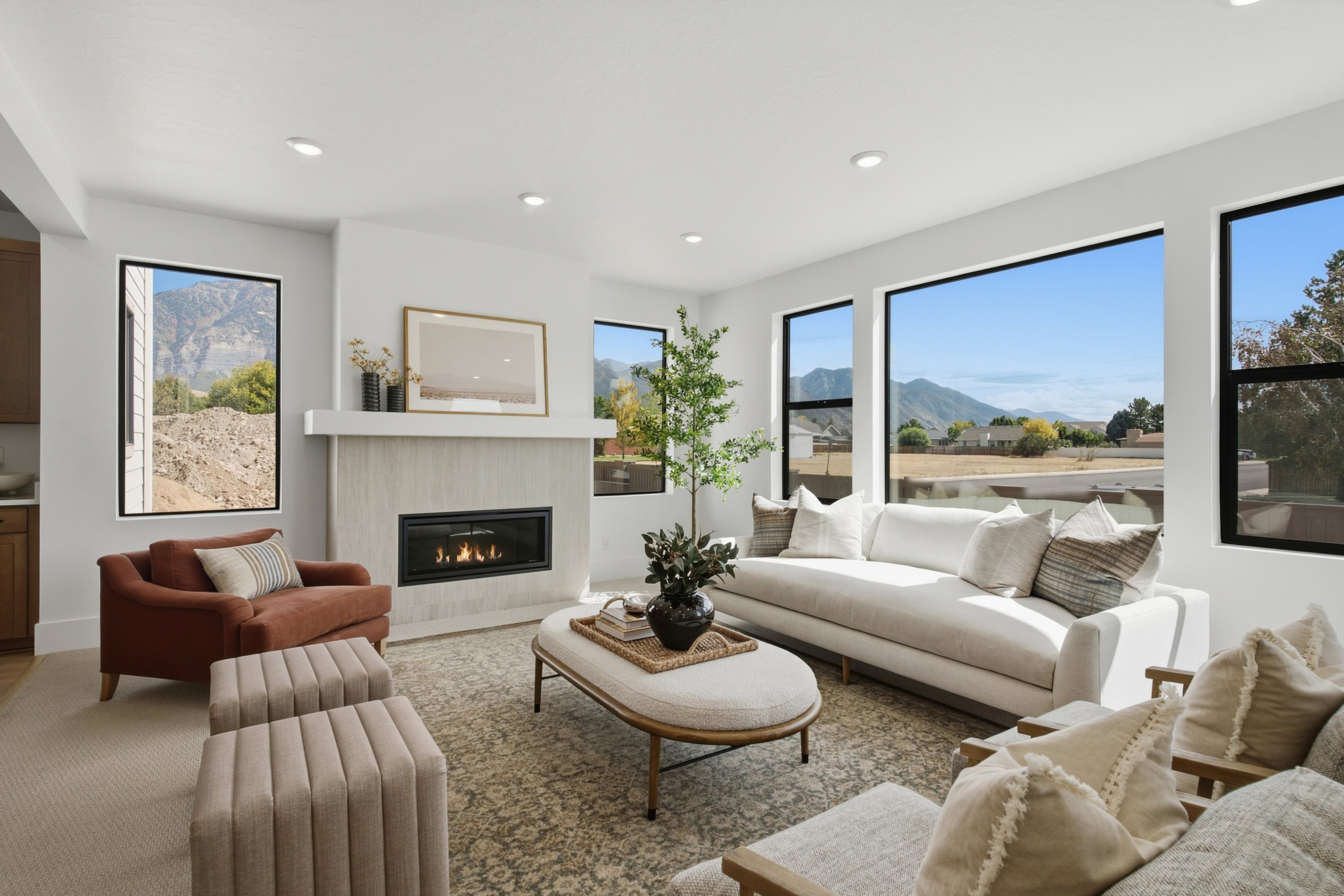 A modern living room with a white sofa, rust-colored armchair, and oval coffee table sits before a fireplace, with large windows revealing a mountainous landscape in the background.