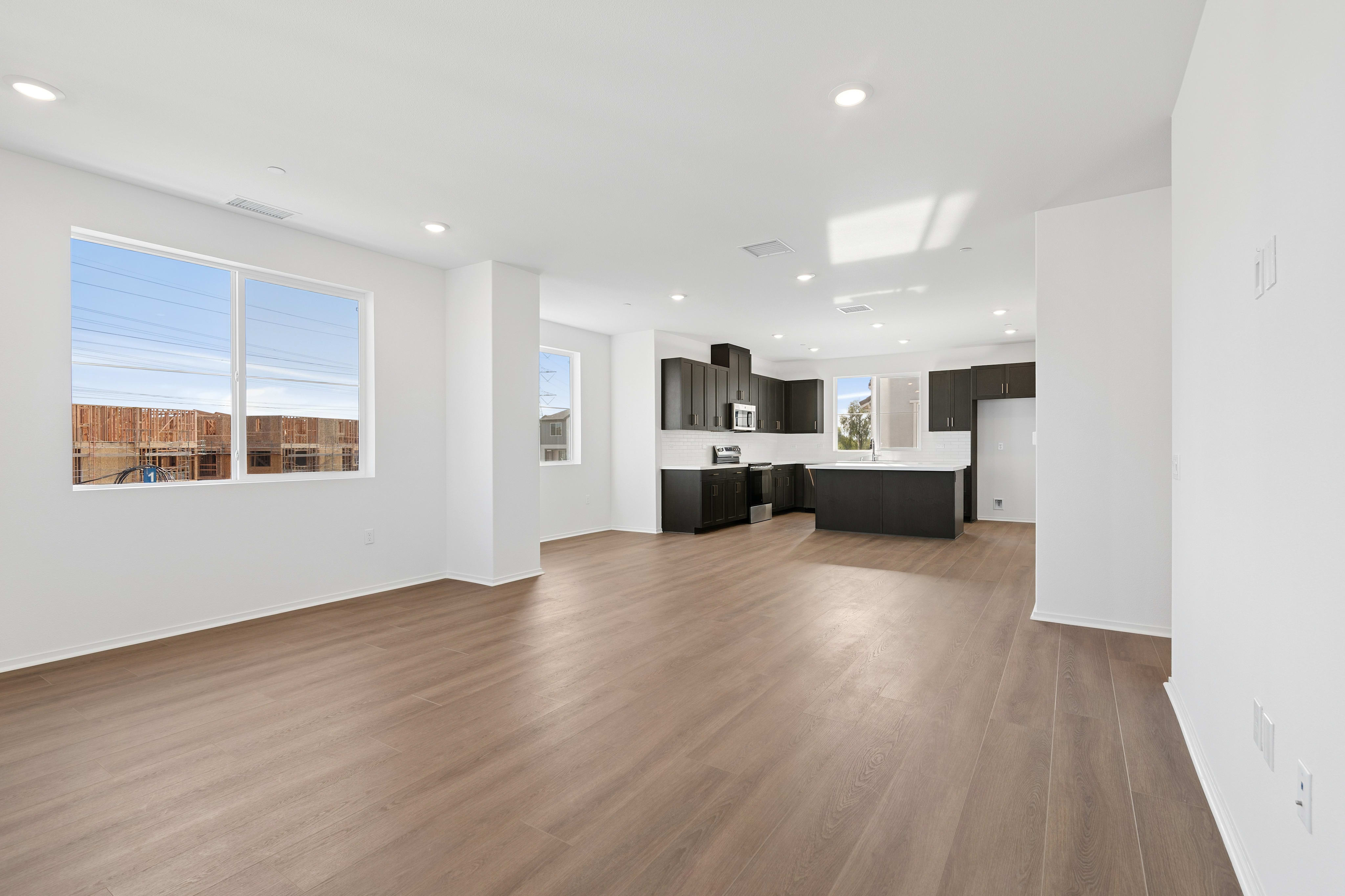 An empty room with light wood flooring leads into a modern kitchen with dark cabinetry and white countertops in the background.