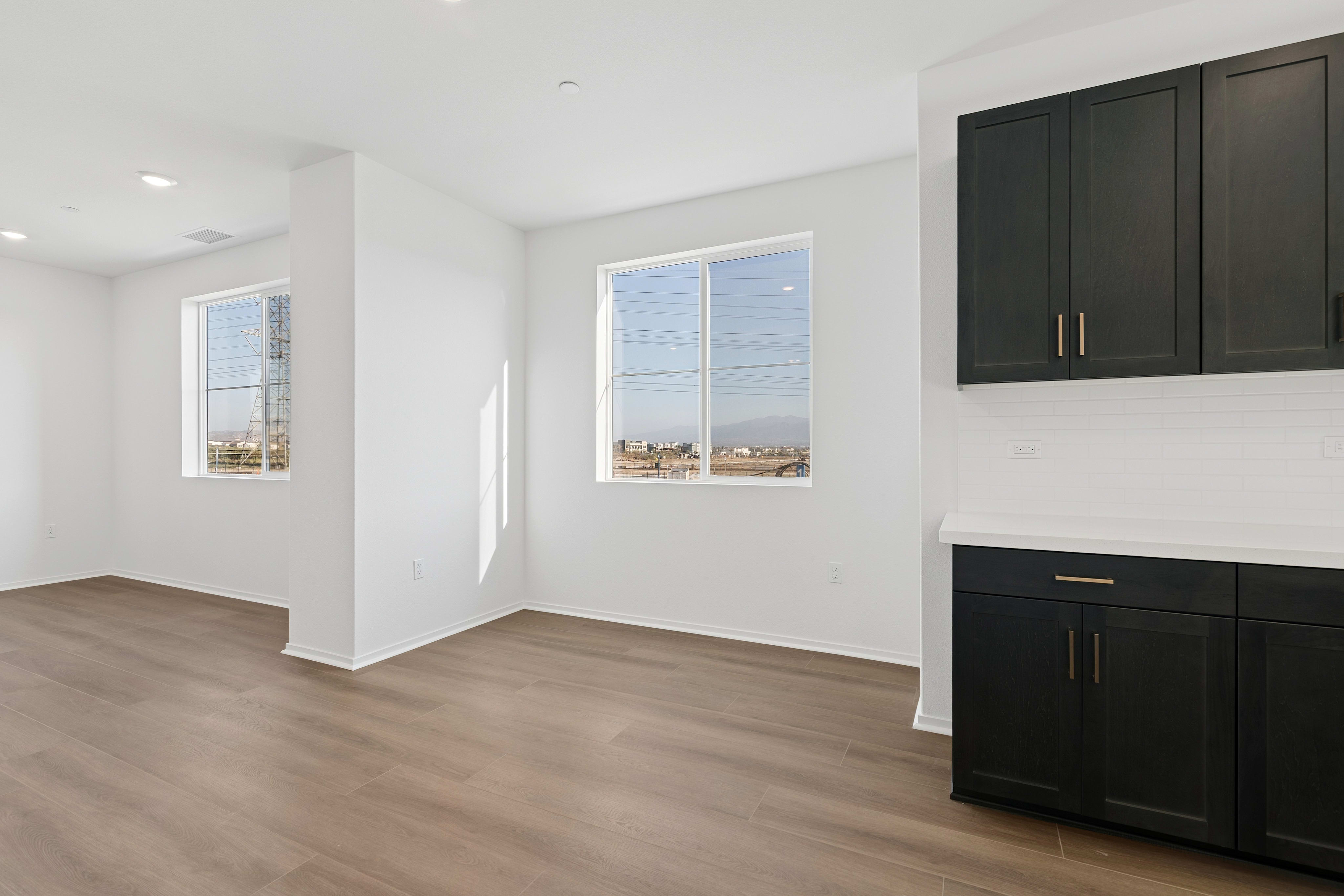 An empty room with light wood flooring and white walls features dark cabinetry in the foreground and two windows looking out onto a landscape in the background.