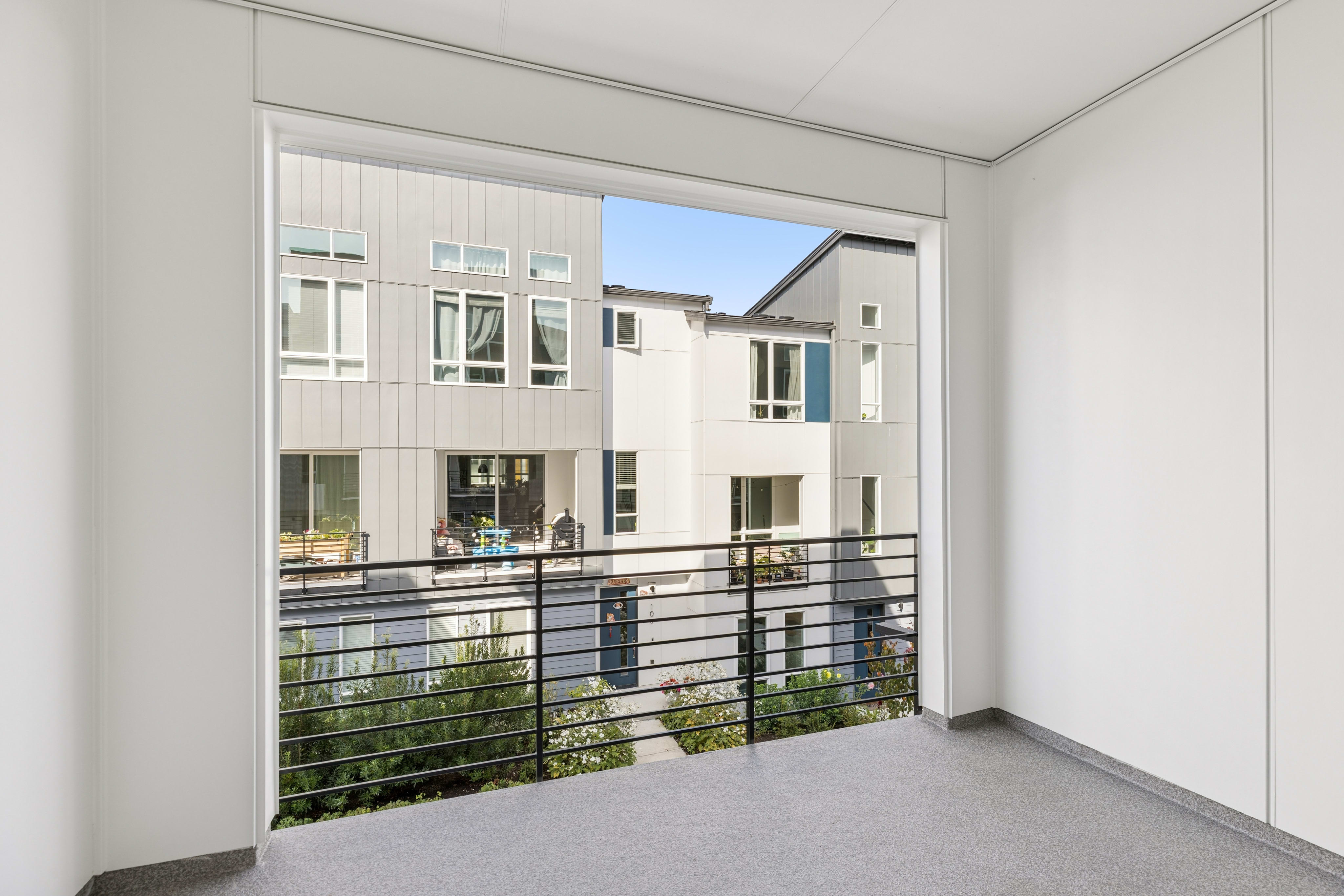 A grey-carpeted balcony with a black metal railing overlooks modern, multi-story white and grey apartment buildings under a clear blue sky.