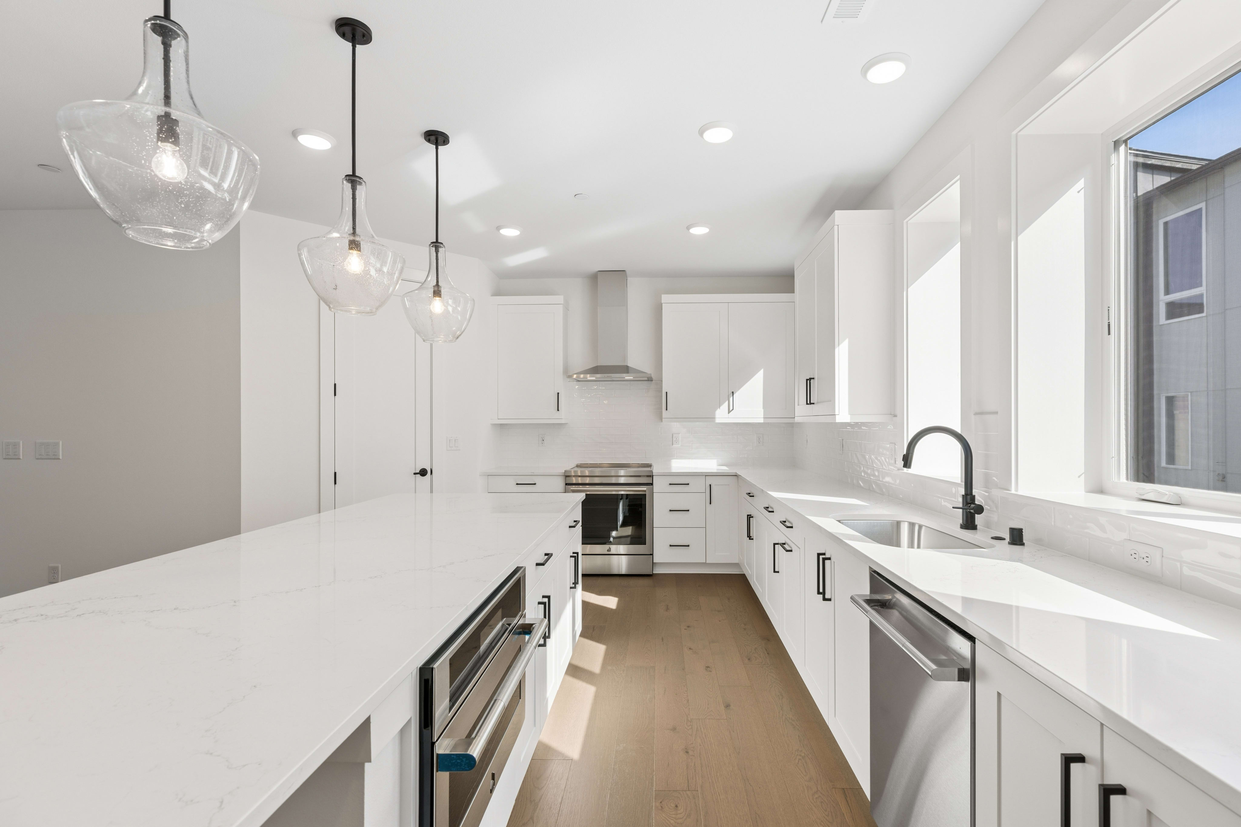 A white marble kitchen island with modern pendant lights sits in the foreground of a bright, all-white kitchen with stainless steel appliances and light wood flooring.