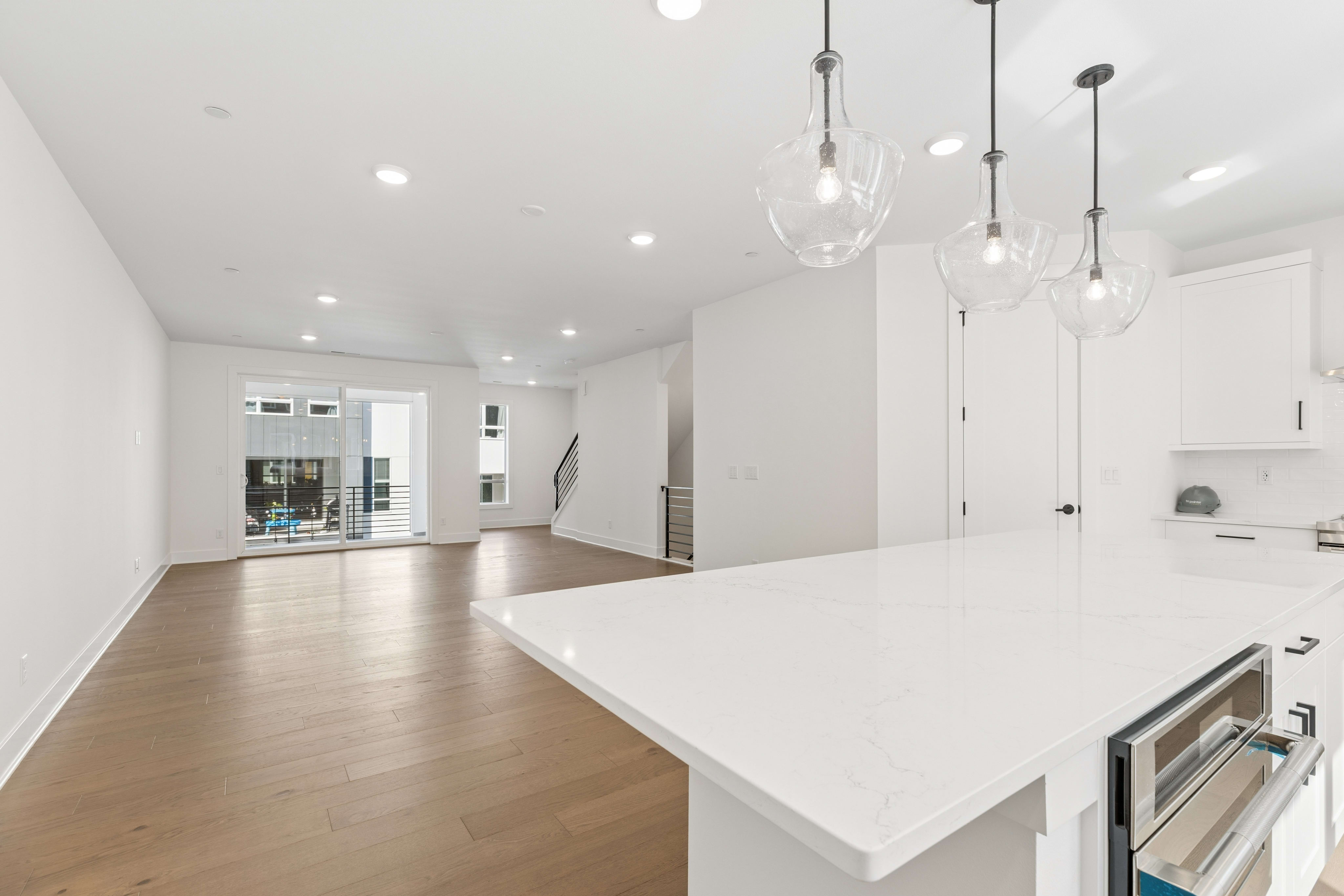 A white quartz kitchen island with three glass pendant lights sits in the foreground of a bright, open-concept living area with light wood floors and a sliding glass door in the background.