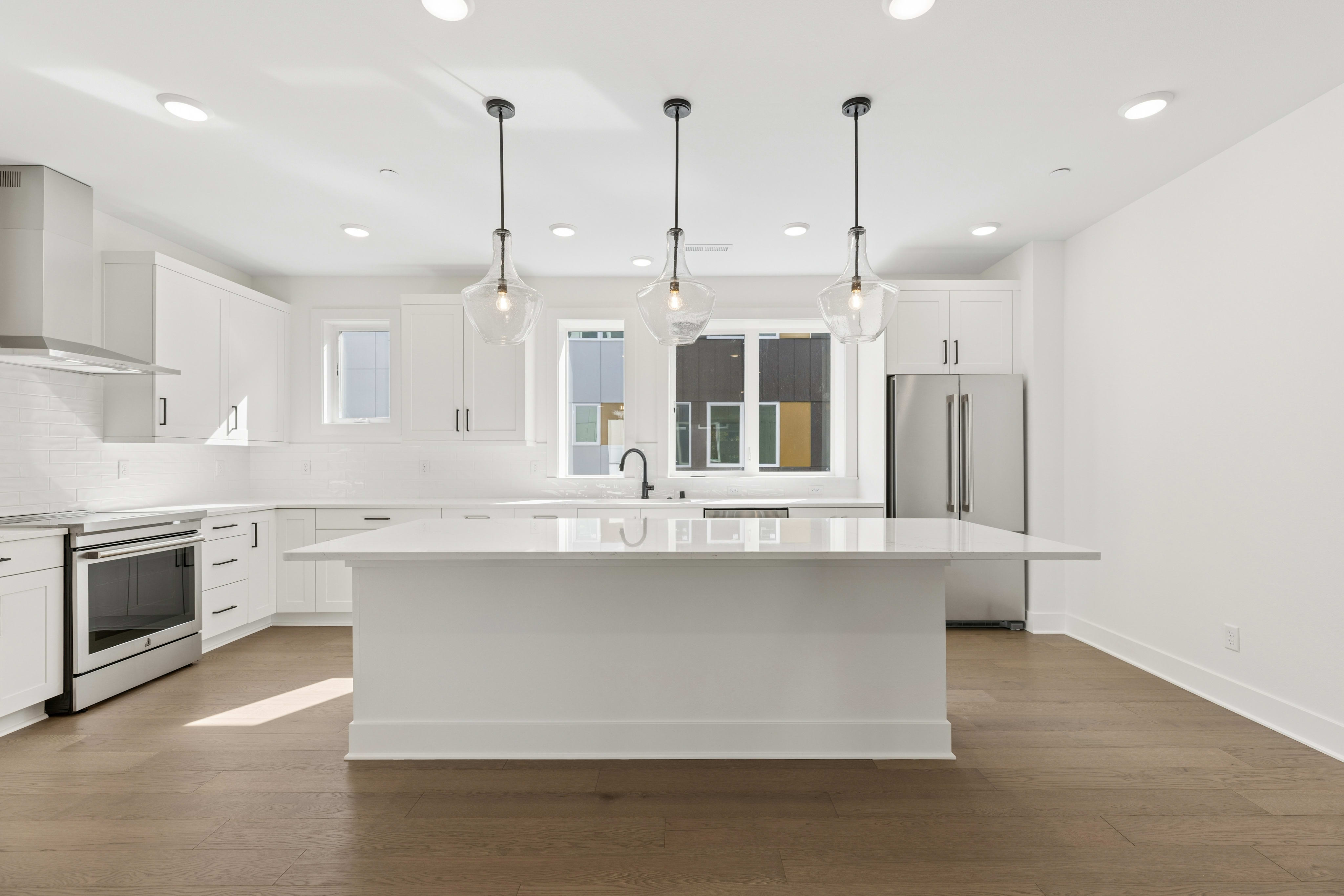 A large white kitchen island sits in the foreground of a bright, modern kitchen with white cabinetry and stainless steel appliances in the background.