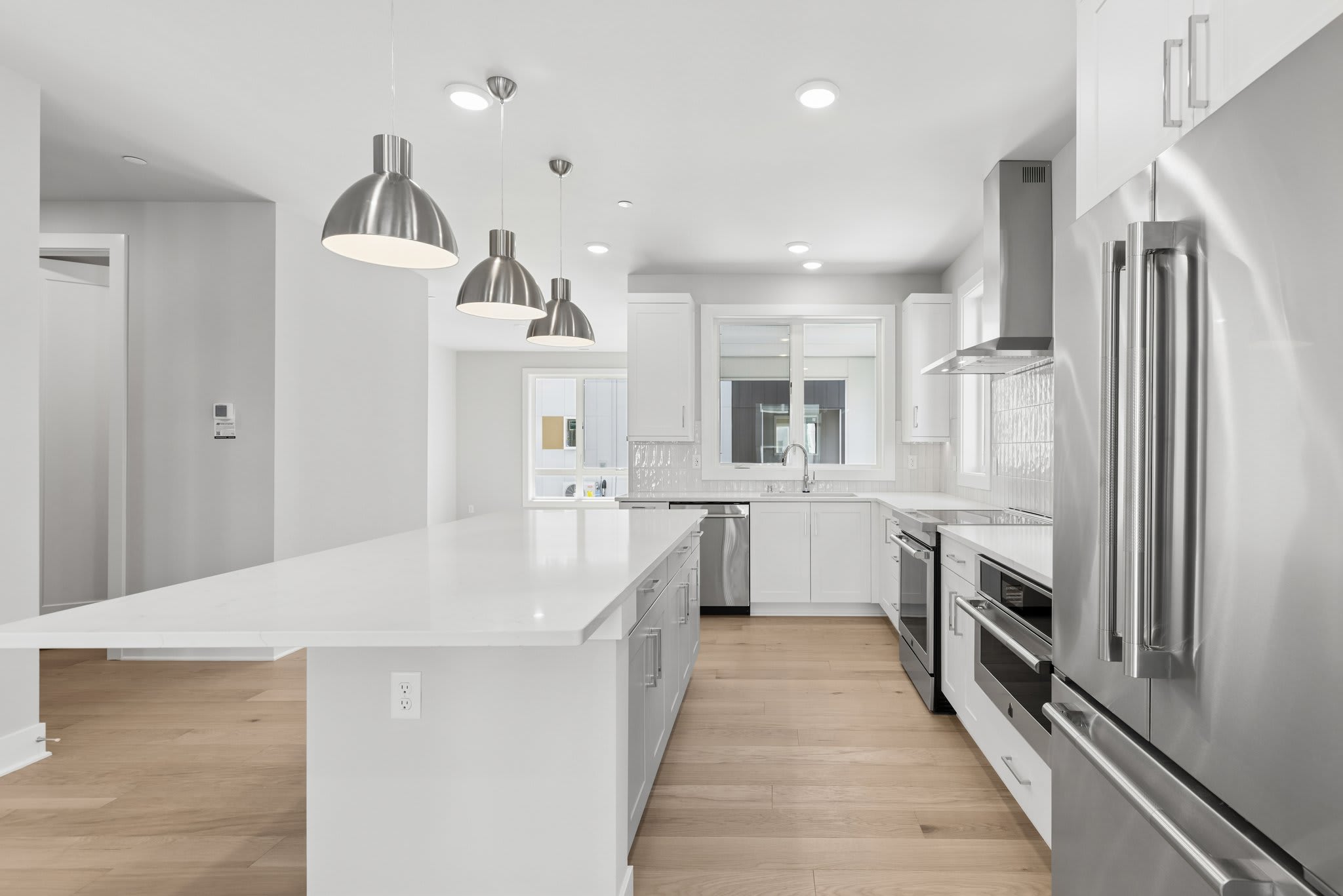 A white kitchen island with three silver pendant lights sits in the foreground of a modern, bright white kitchen with stainless steel appliances.