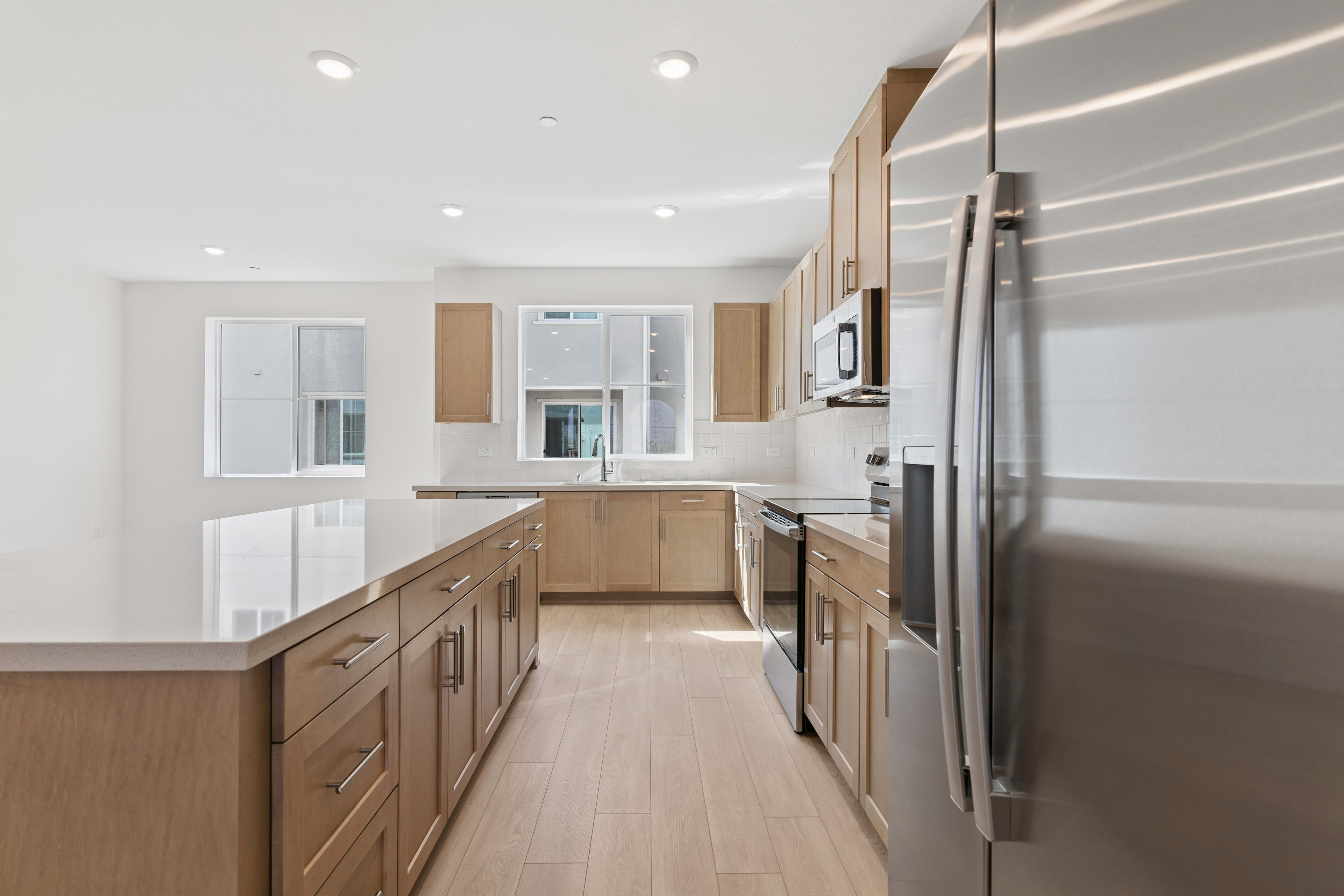 A stainless steel refrigerator stands in the foreground of a modern kitchen with light wood cabinetry and white countertops.