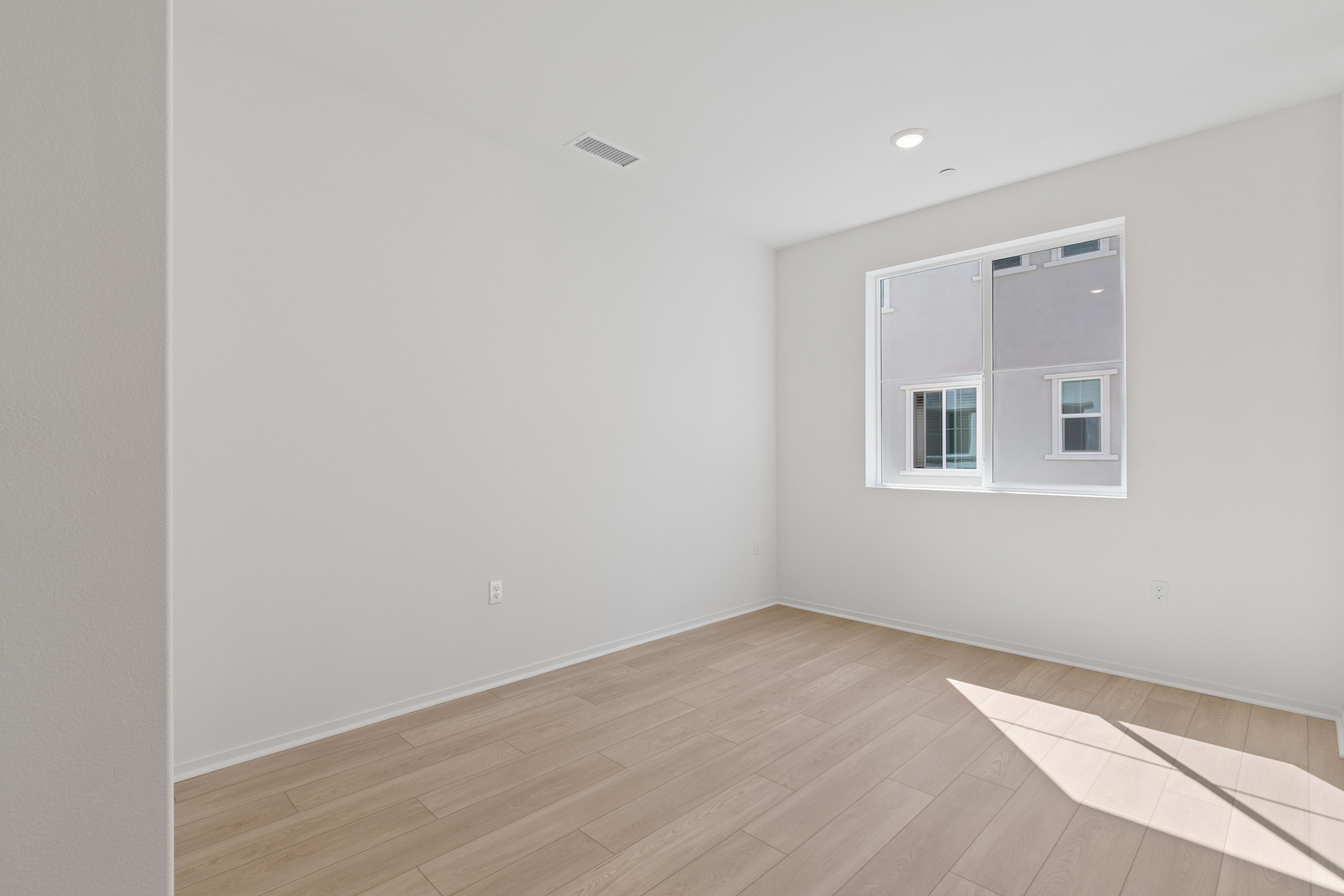 An empty room with light wood flooring in the foreground and white walls with a window in the background.
