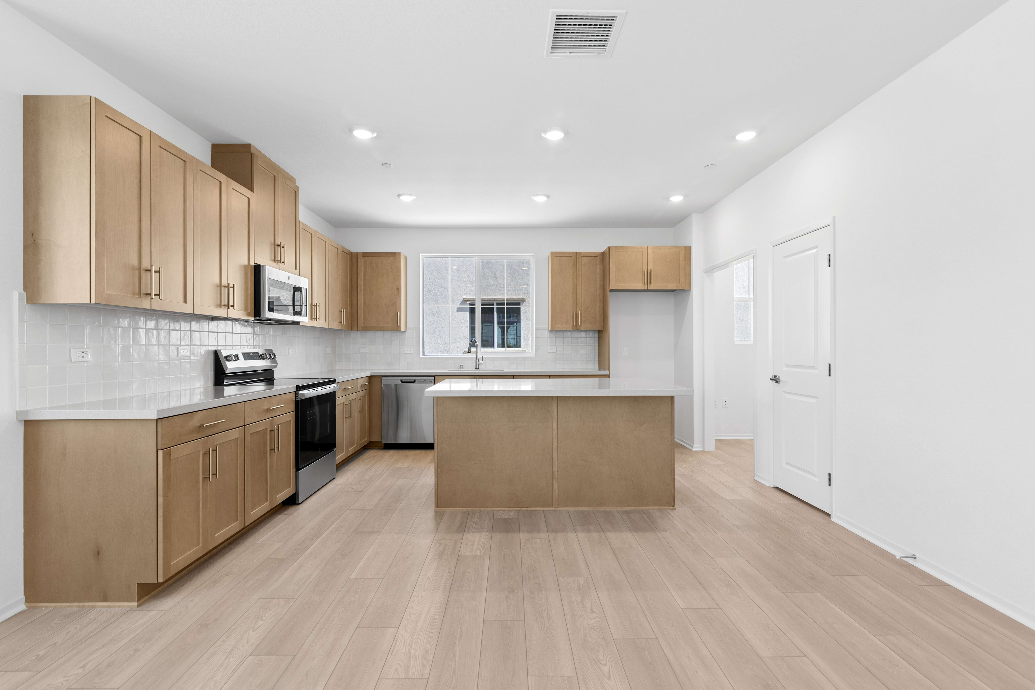 A modern kitchen features light wood cabinetry and white countertops in the foreground, with a white door and bright walls in the background.