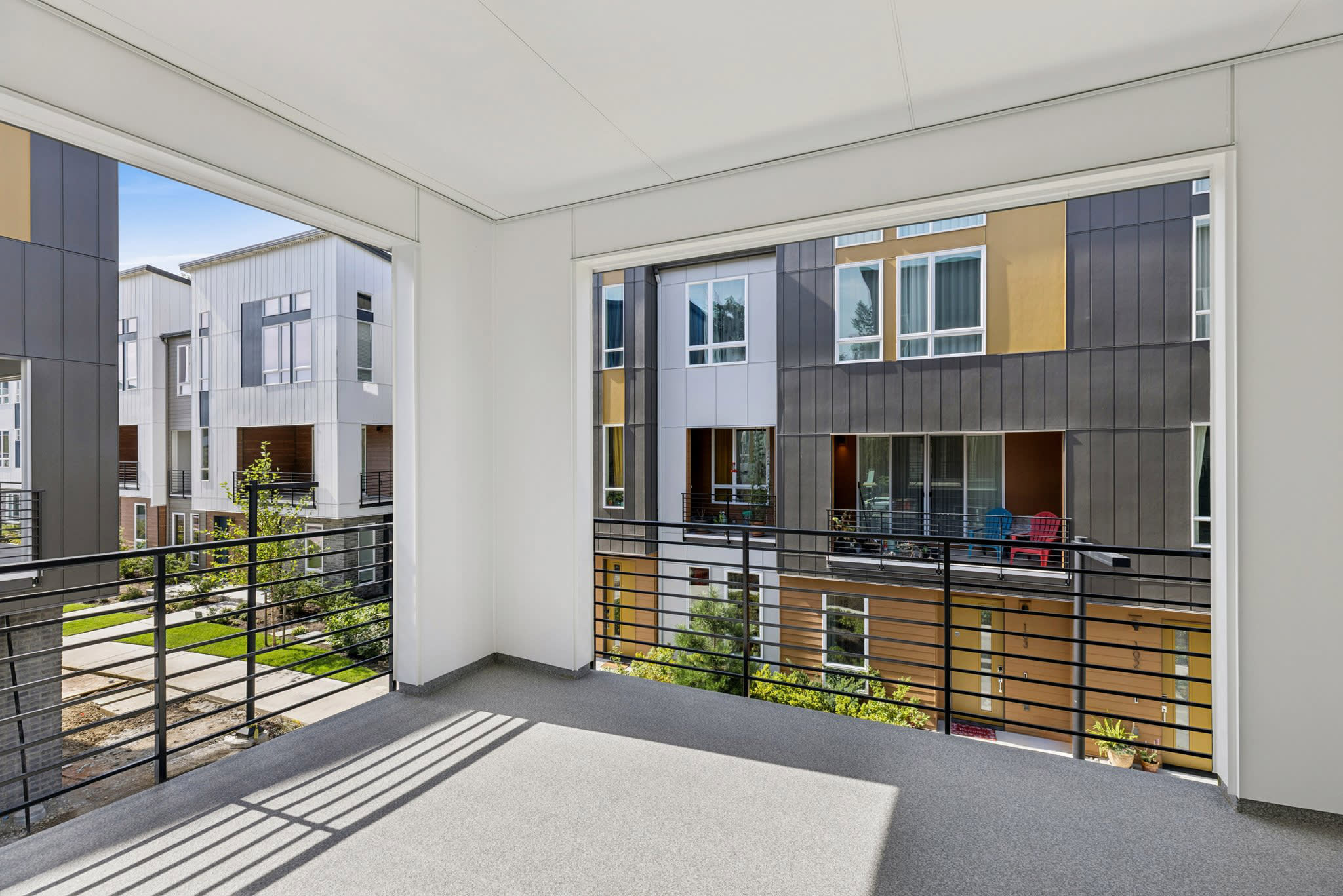 A grey carpeted balcony with black metal railings overlooks a modern multi-story apartment complex with grey and tan siding.