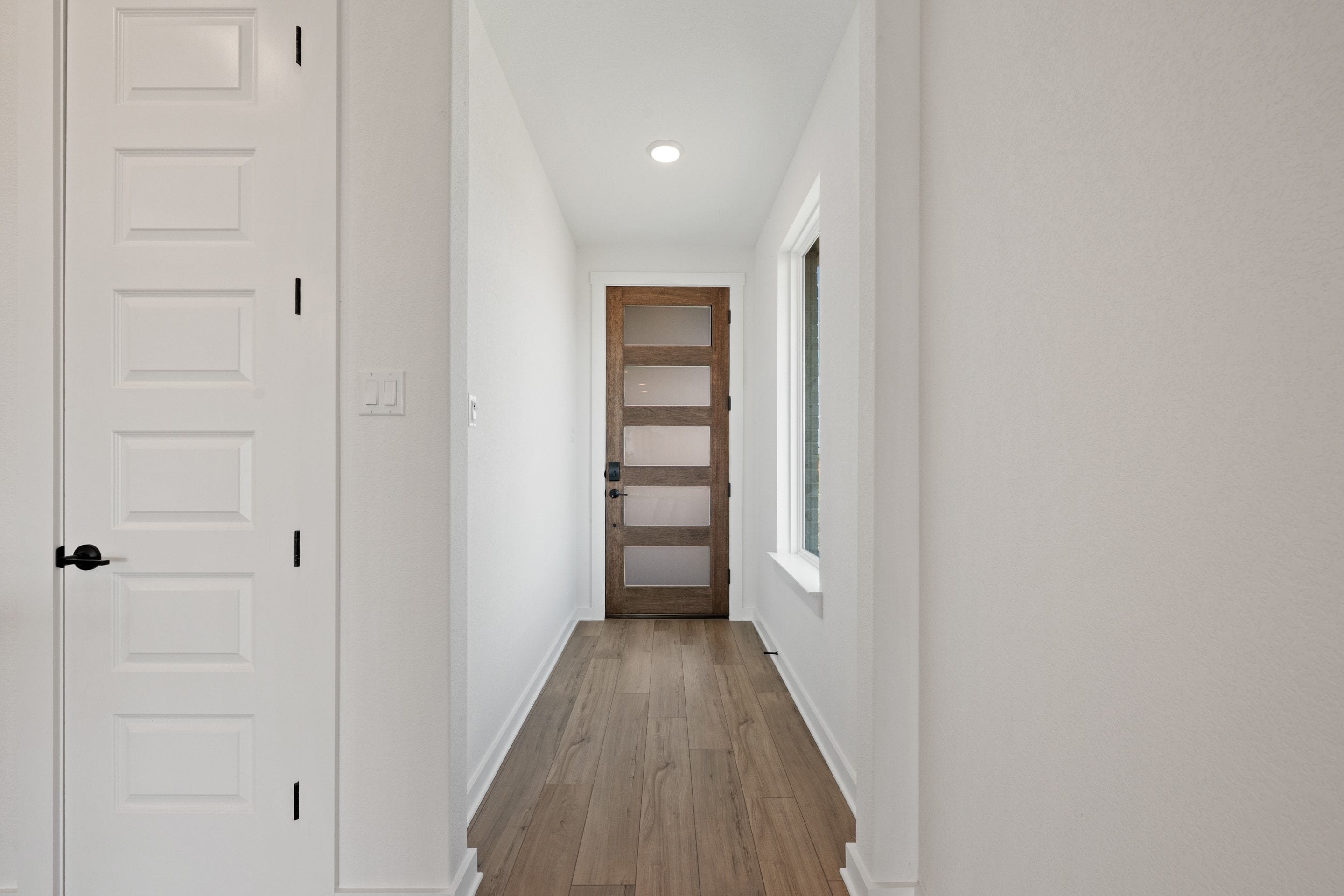 A white paneled door stands in the foreground of a bright hallway leading to a wooden glass-paneled door in the background.