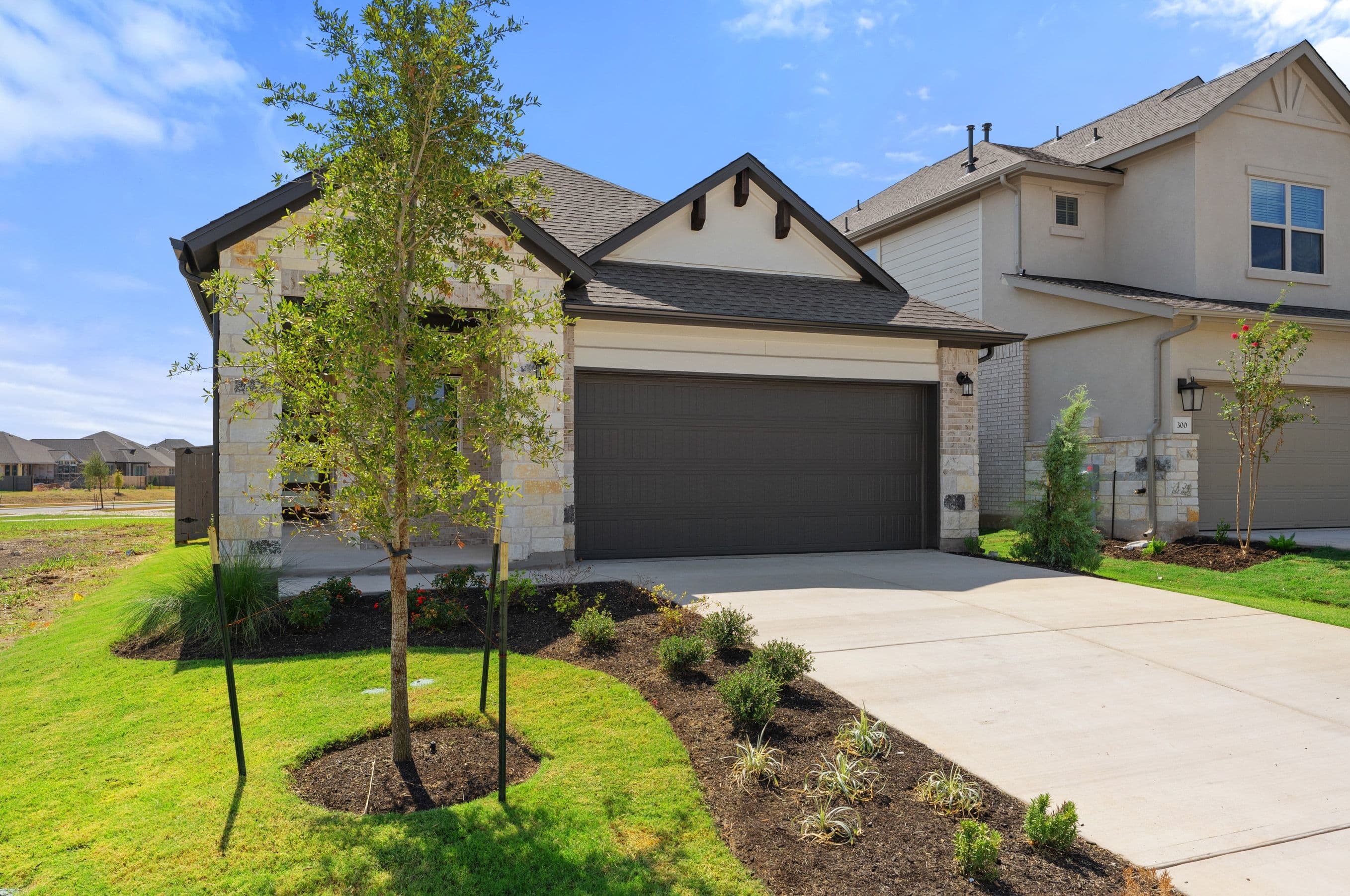 A young tree and manicured lawn stand in the foreground of a modern single-story home with a dark garage door, set against a clear blue sky and a neighboring house.