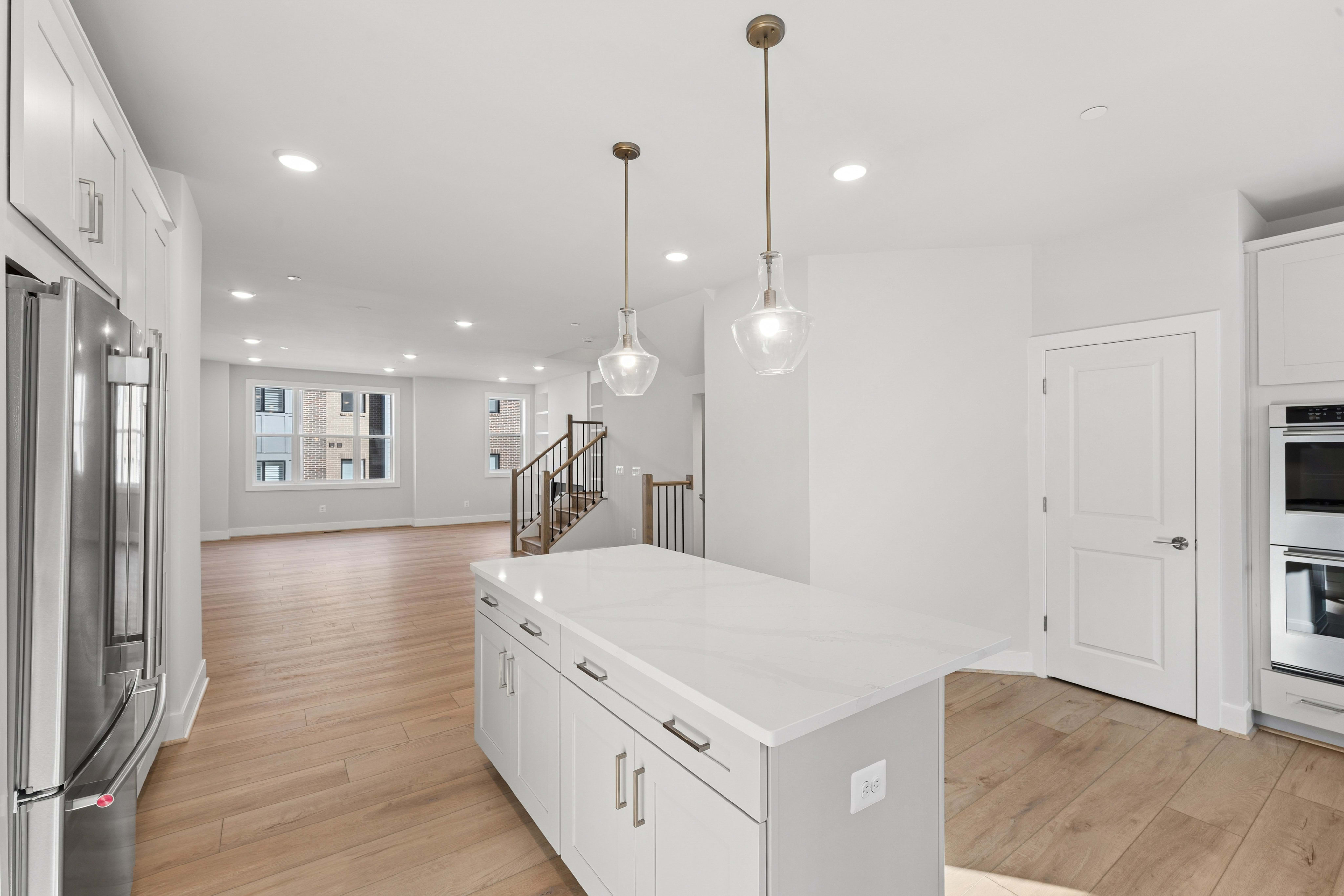 A white kitchen island with pendant lights sits in the foreground of a bright, open-concept living space with light wood flooring and a staircase in the background.