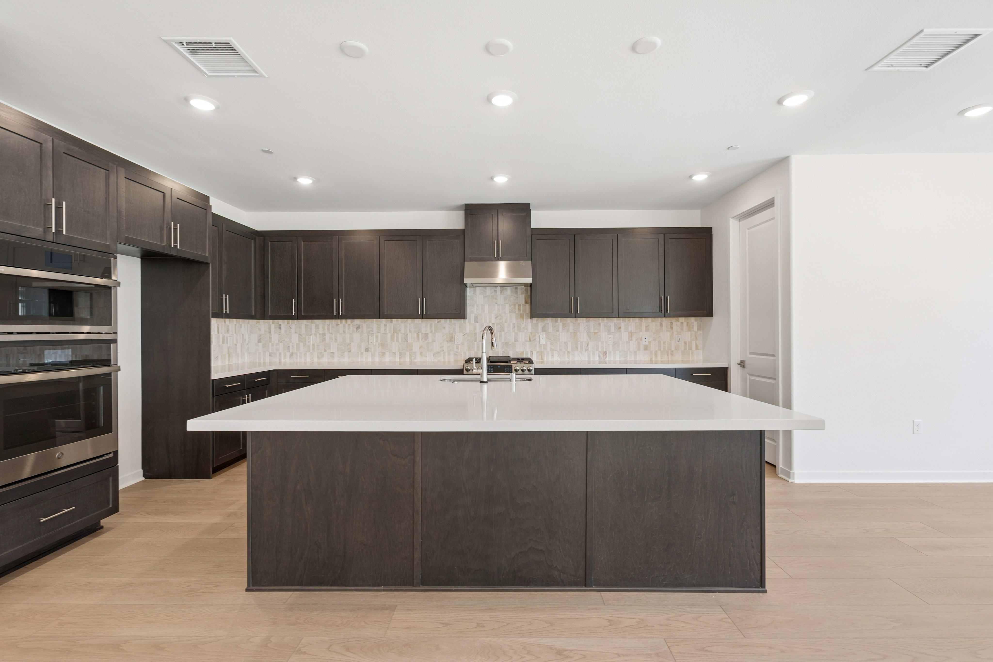 A large white kitchen island sits in the foreground of a modern kitchen featuring dark wood cabinetry and a tiled backsplash.