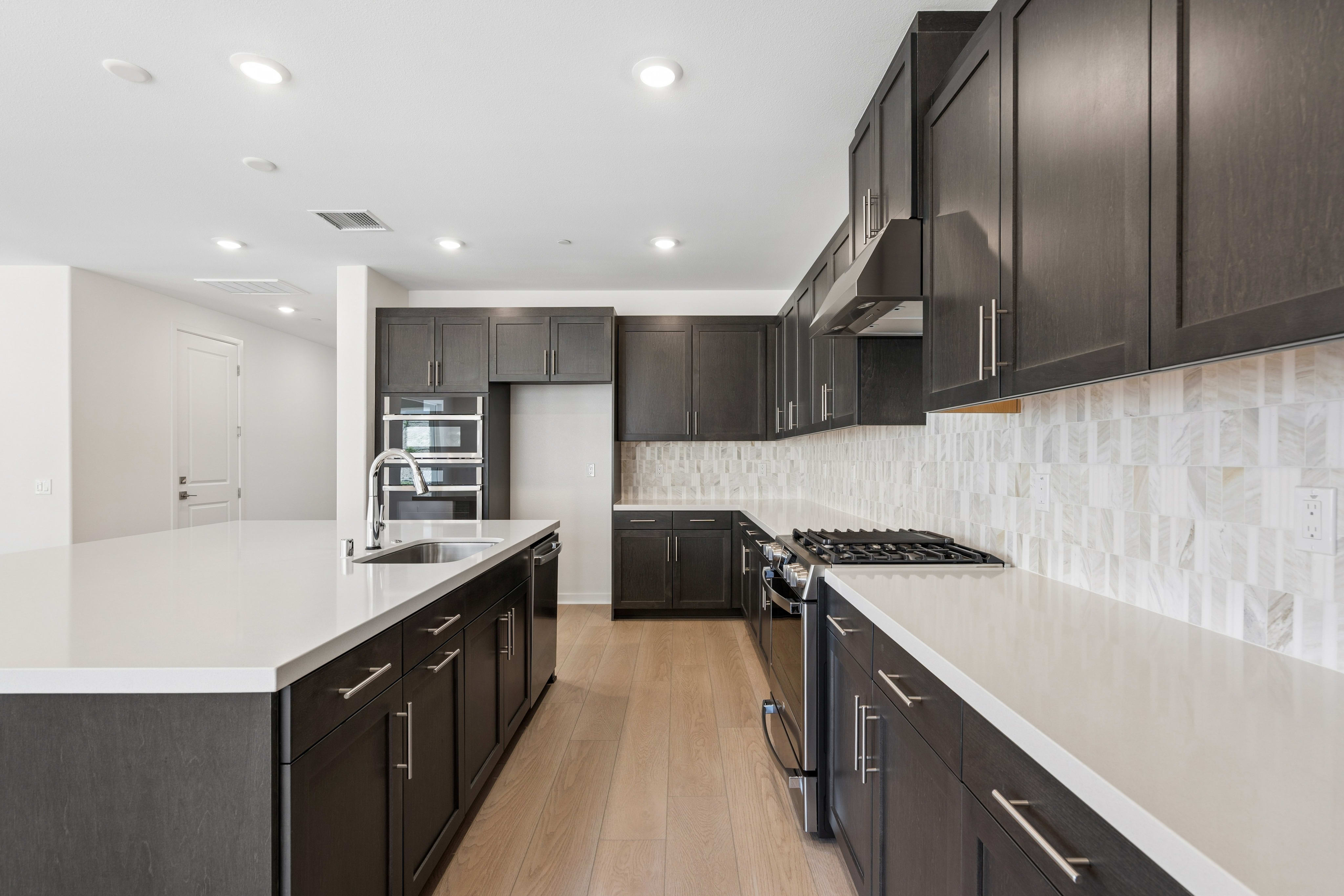 A modern kitchen features a white quartz island and dark wood cabinetry in the foreground, with a matching cabinet set and a white tiled backsplash in the background.