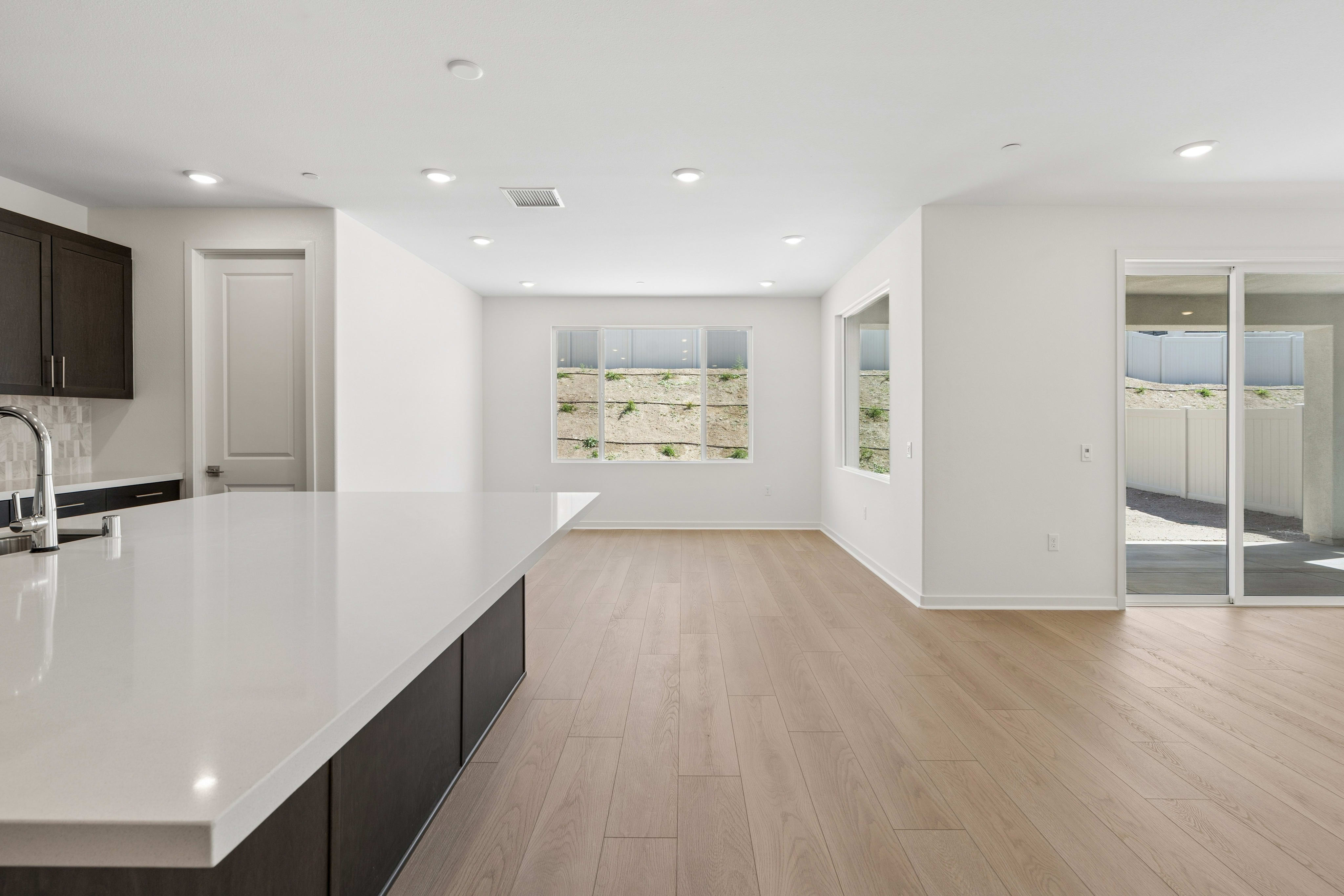 A white kitchen countertop and dark cabinetry occupy the foreground of a bright, open-concept living space with light wood floors and large windows looking out onto a sandy landscape.