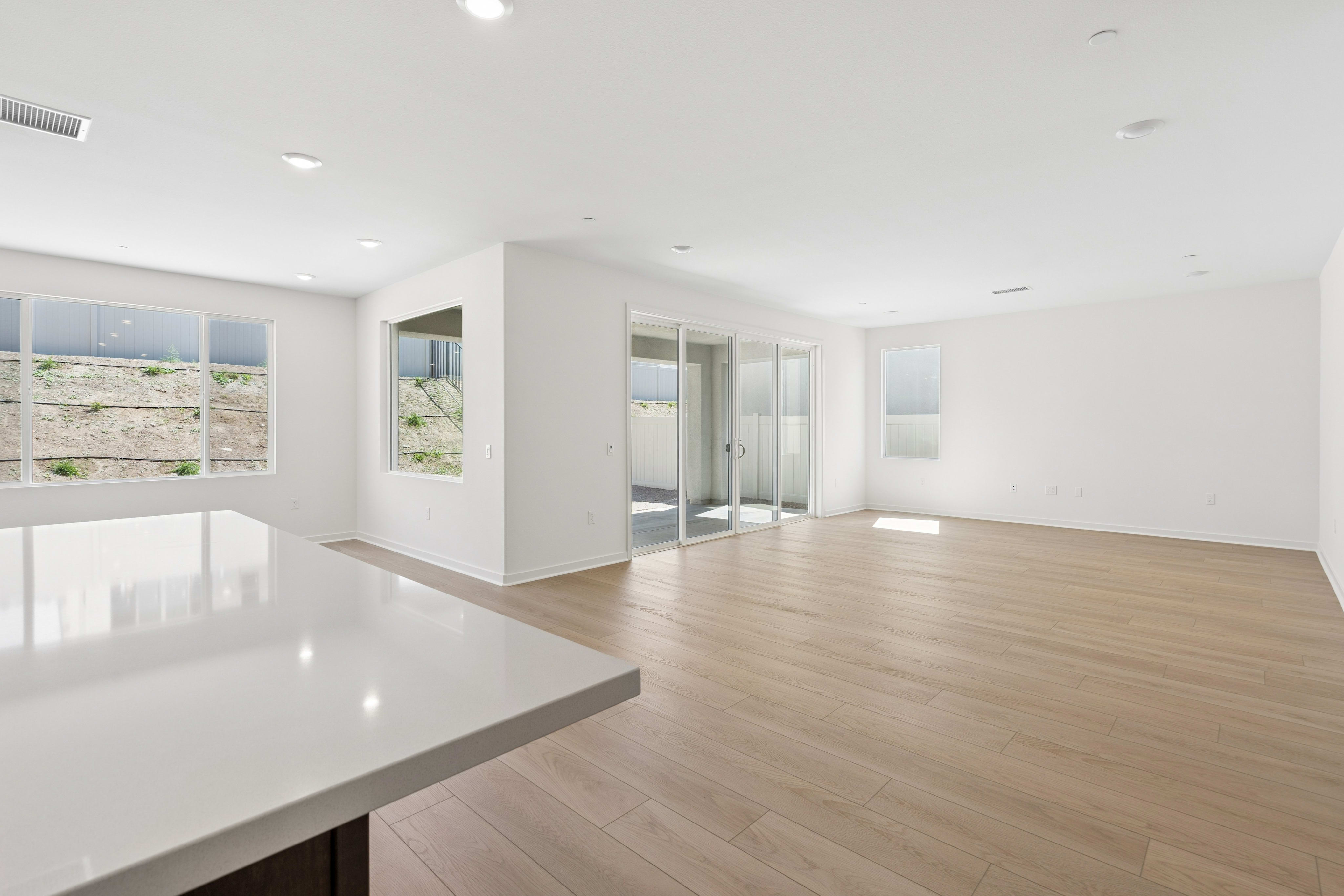 A white kitchen countertop sits in the foreground of an empty, open-concept living space with light wood floors and large windows looking out onto a hillside.