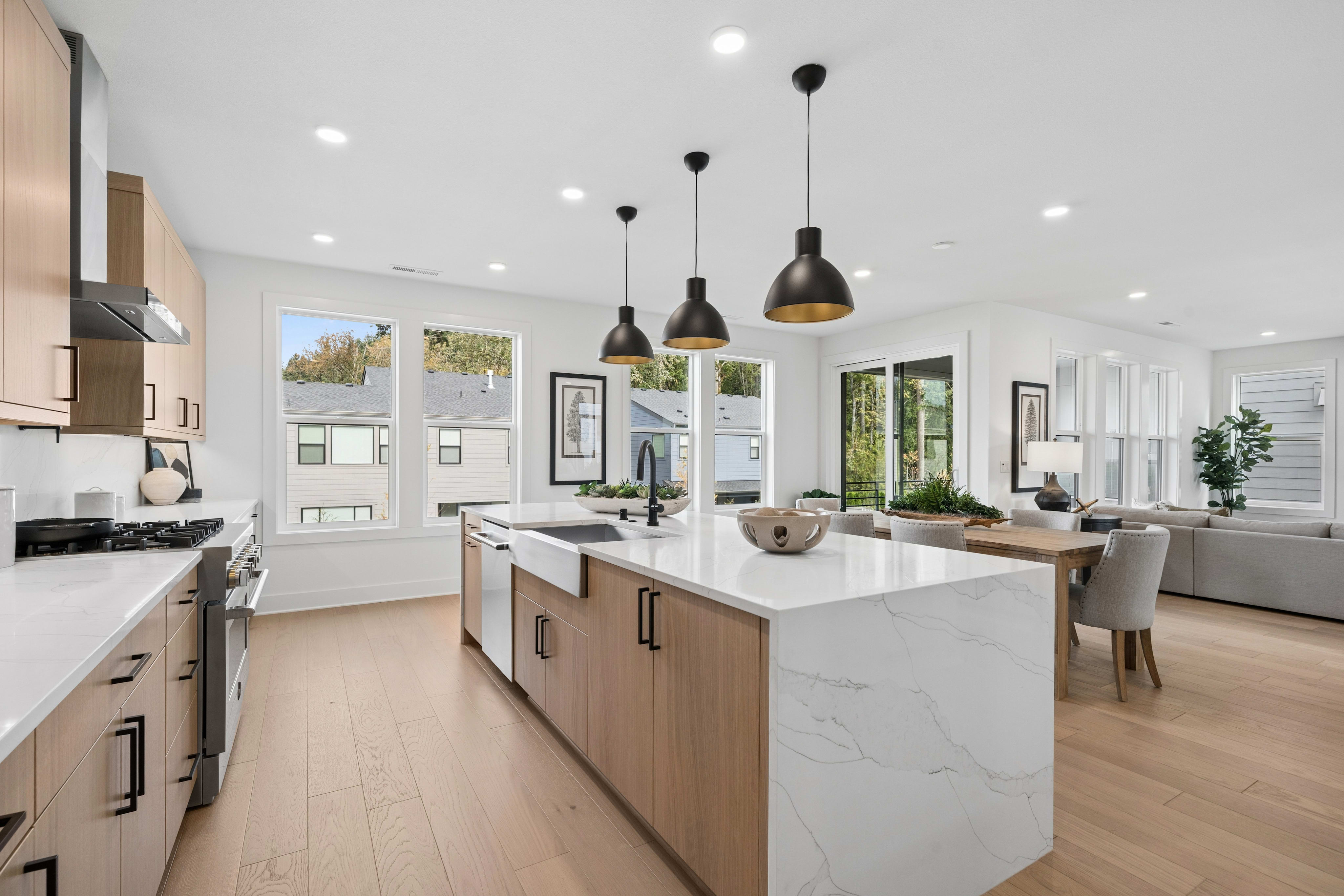 A modern kitchen with a white marble island and light wood cabinetry sits in the foreground, opening into a bright living area with a dining table and grey sofa in the background.