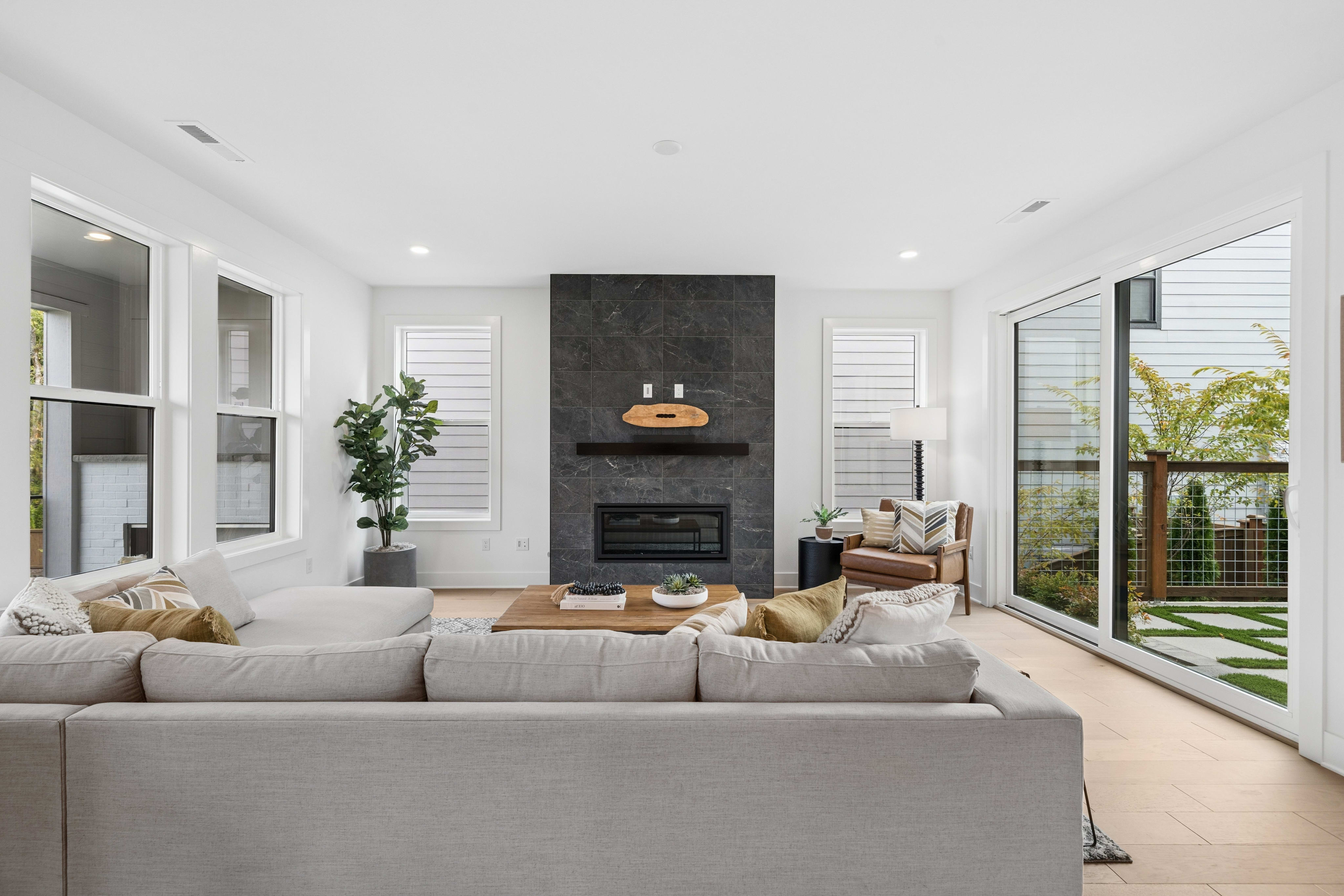 A light gray sectional sofa sits in the foreground of a bright living room featuring a dark marble fireplace and a sliding glass door leading to a backyard.