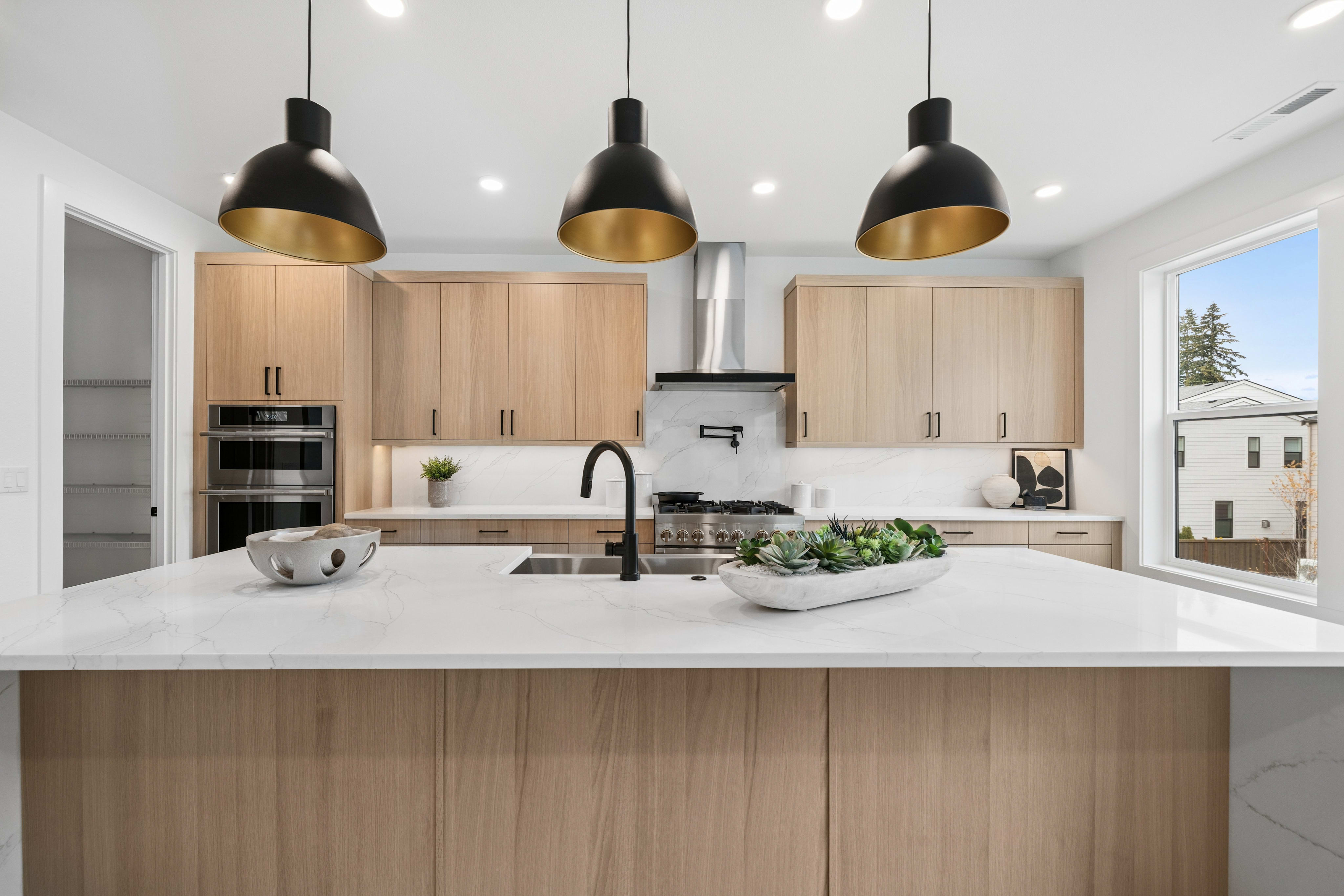 Three black and gold pendant lights hang over a white marble kitchen island in the foreground, with light wood cabinetry and a stainless steel range hood in the background.