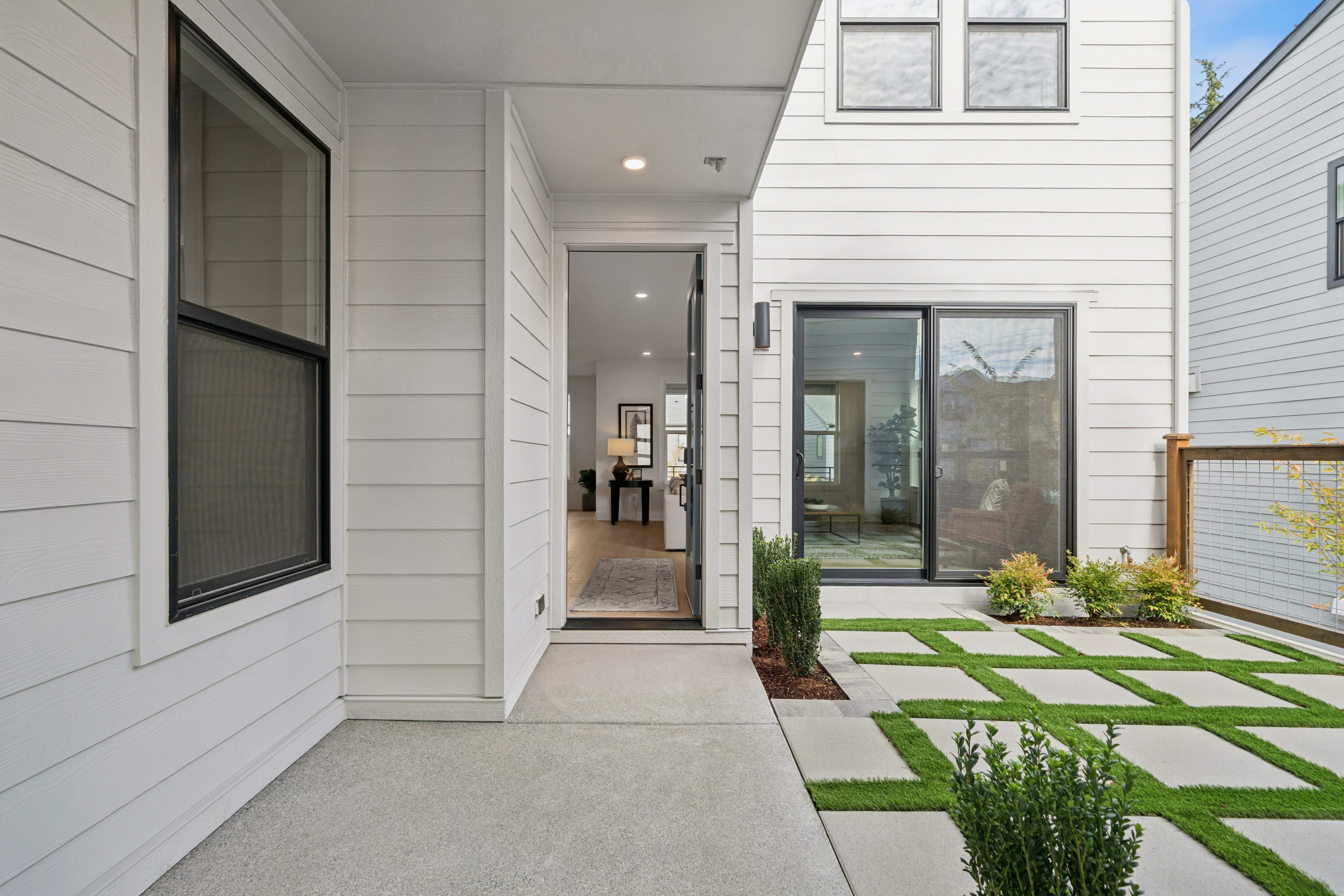 A concrete walkway and geometric grass pavers lead toward the open doorway and sliding glass doors of a modern white house.