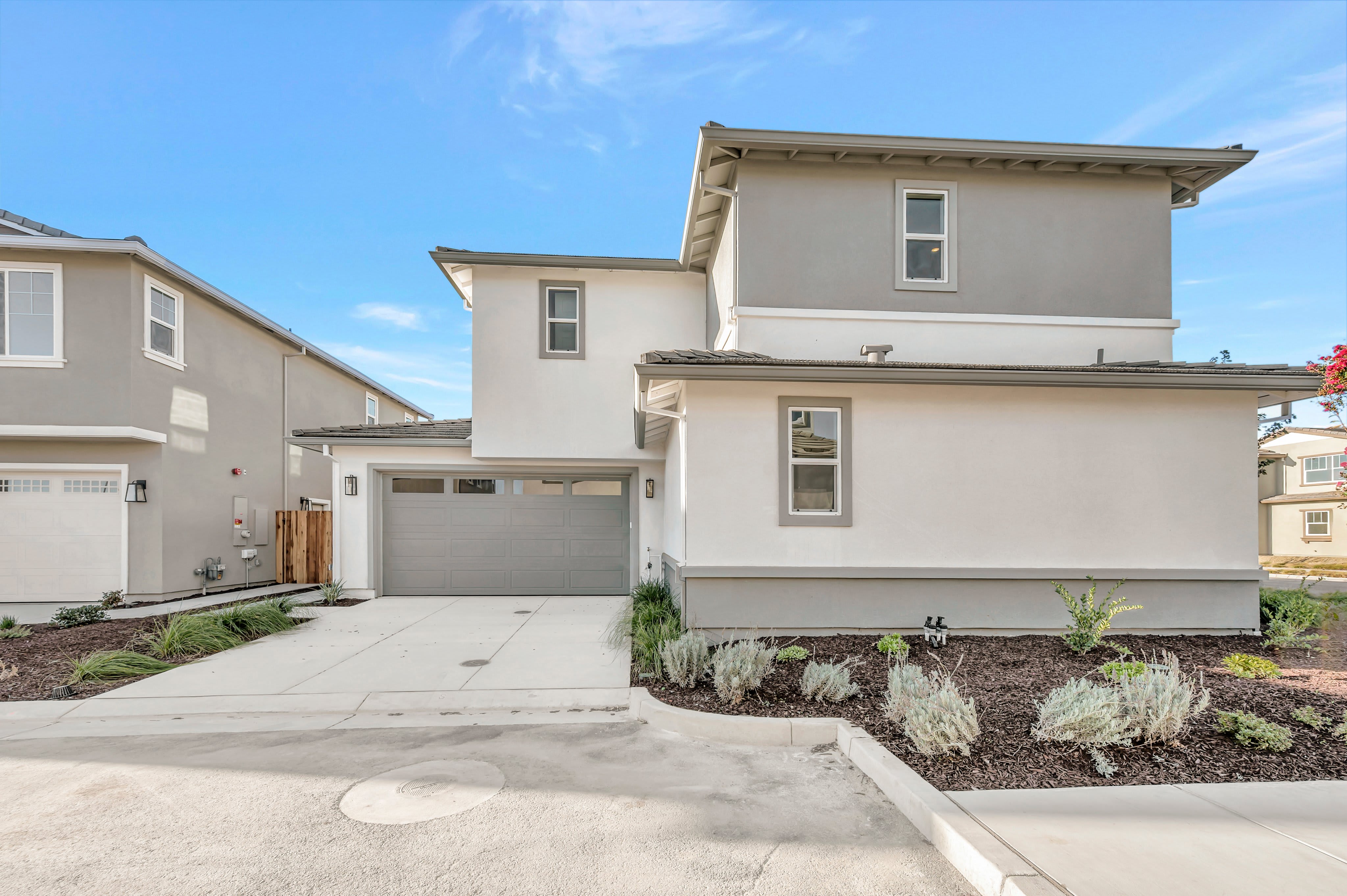 A modern two-story grey house with a concrete driveway and landscaped garden beds sits under a clear blue sky.