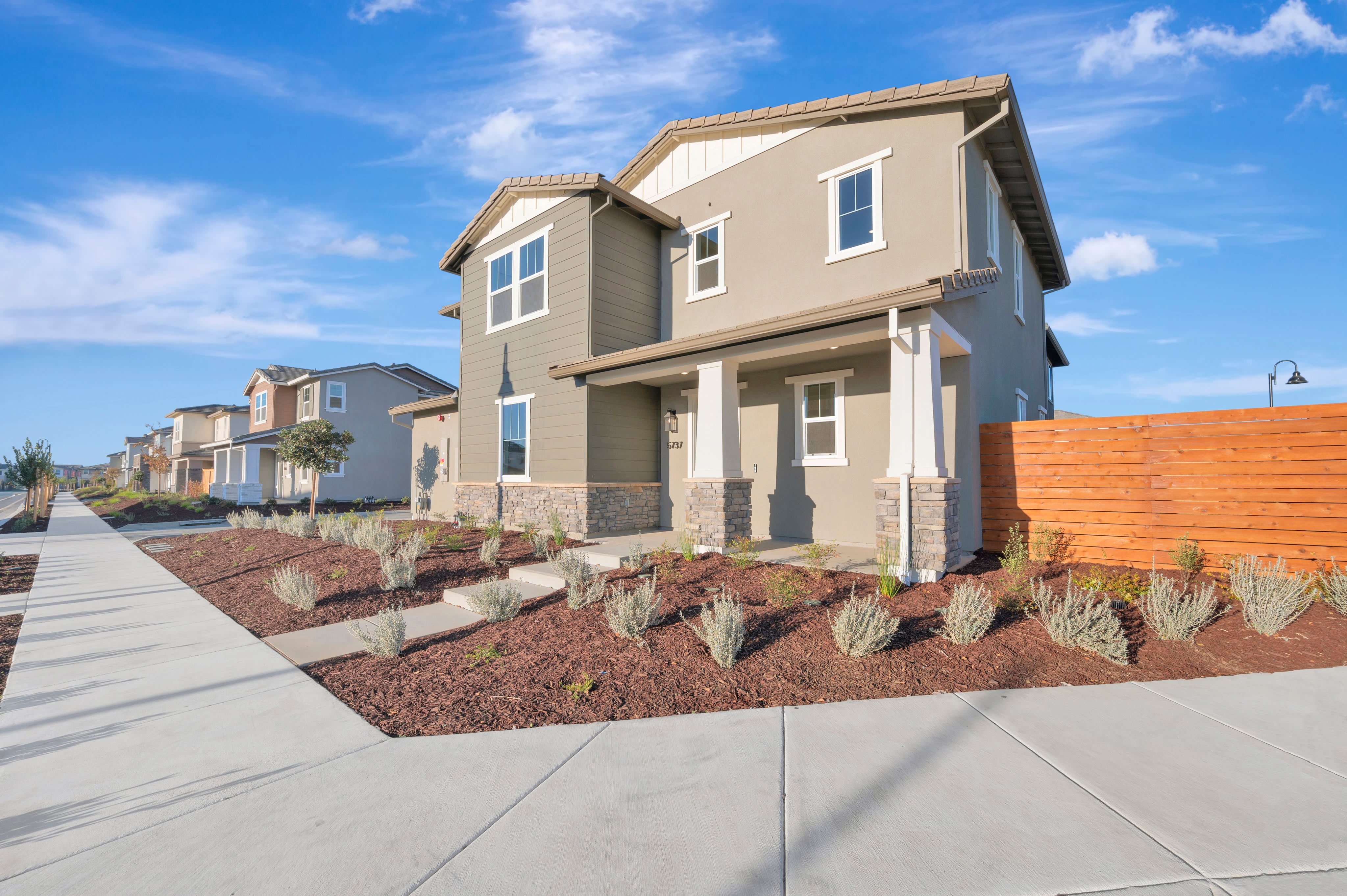 A modern two-story house with a landscaped mulch garden and concrete walkway sits under a bright blue sky with wispy clouds.