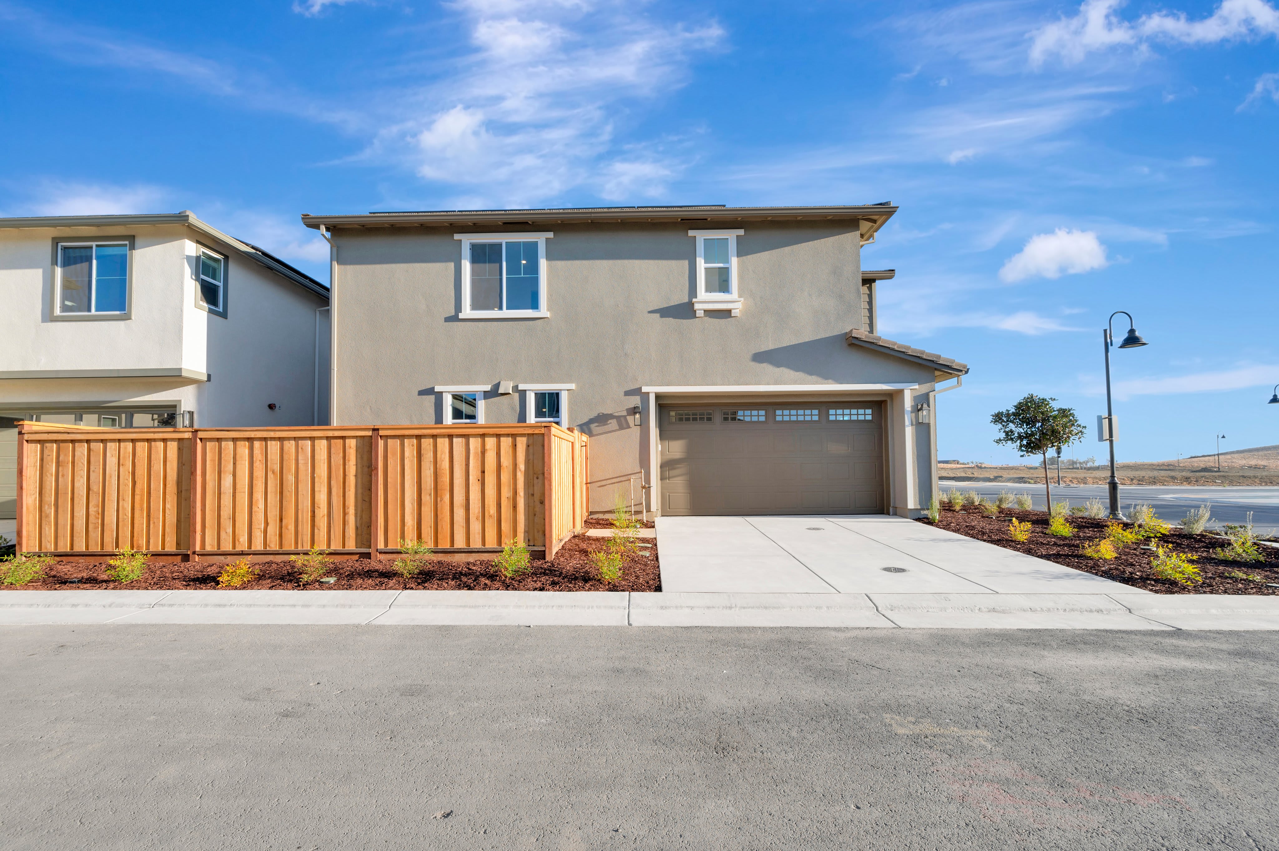 A paved street and wooden fence sit in the foreground of a two-story tan house under a bright blue sky.