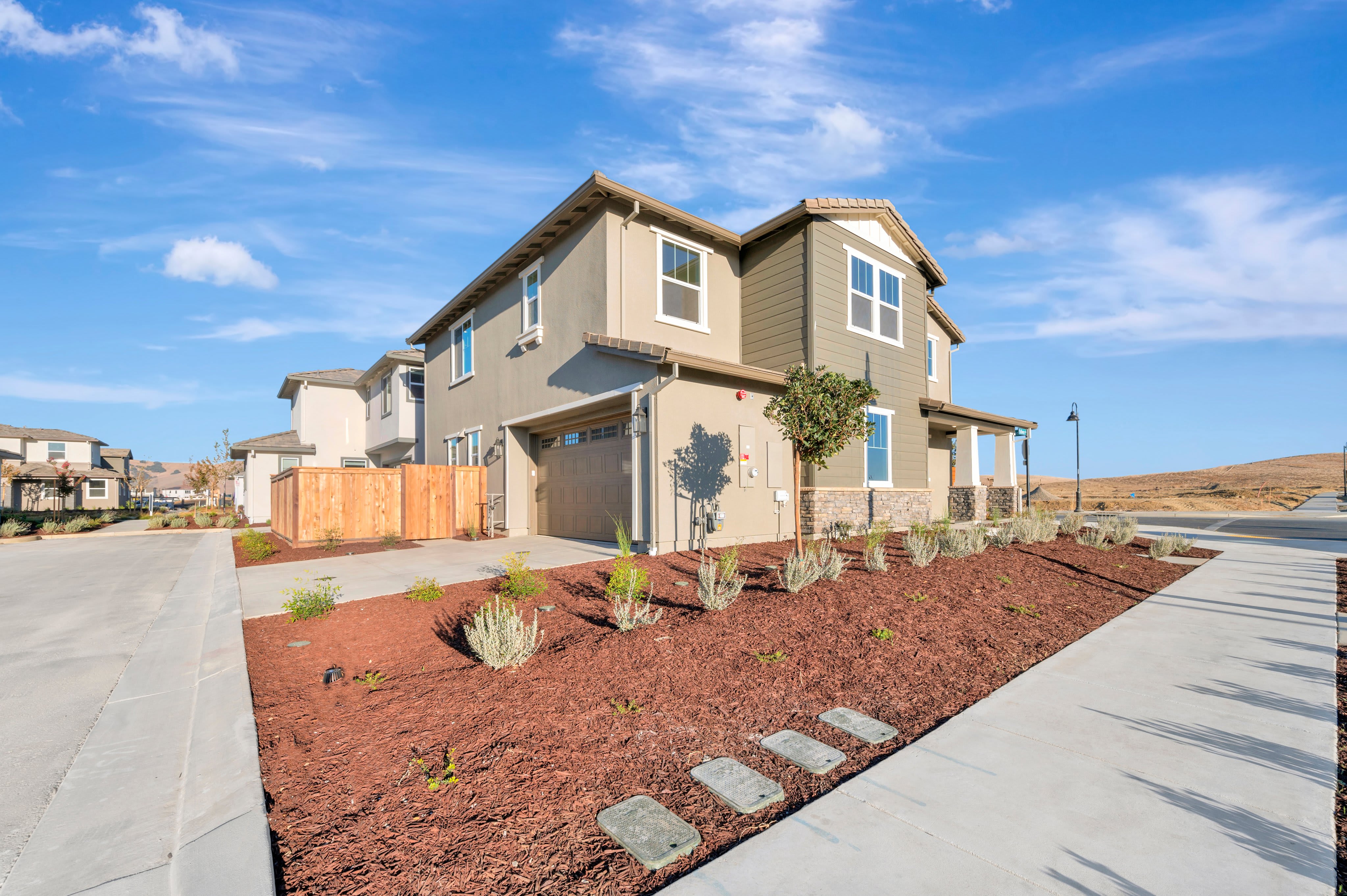 A modern two-story house with a red mulch garden and concrete walkway sits under a bright blue sky with rolling hills in the distance.