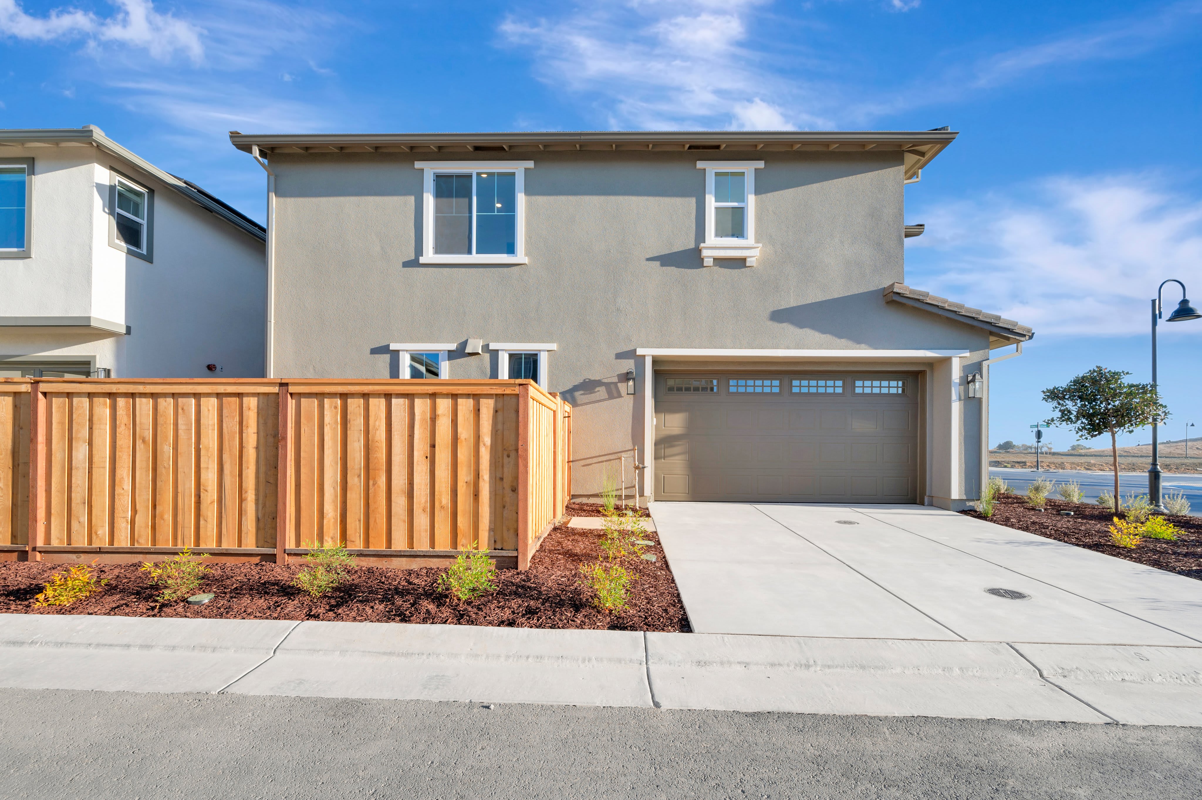 A wooden fence and concrete driveway lead to a two-story grey house under a bright blue sky with wispy clouds.