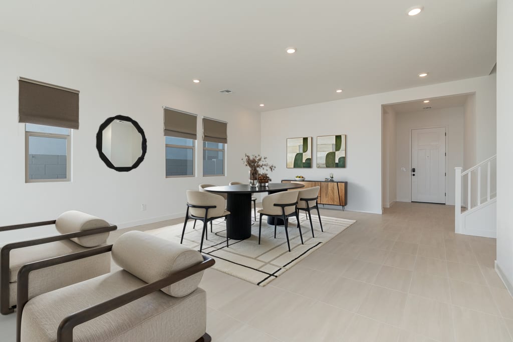 Two modern beige armchairs sit in the foreground of a bright, open living space featuring a round dining table and a white entryway in the background.