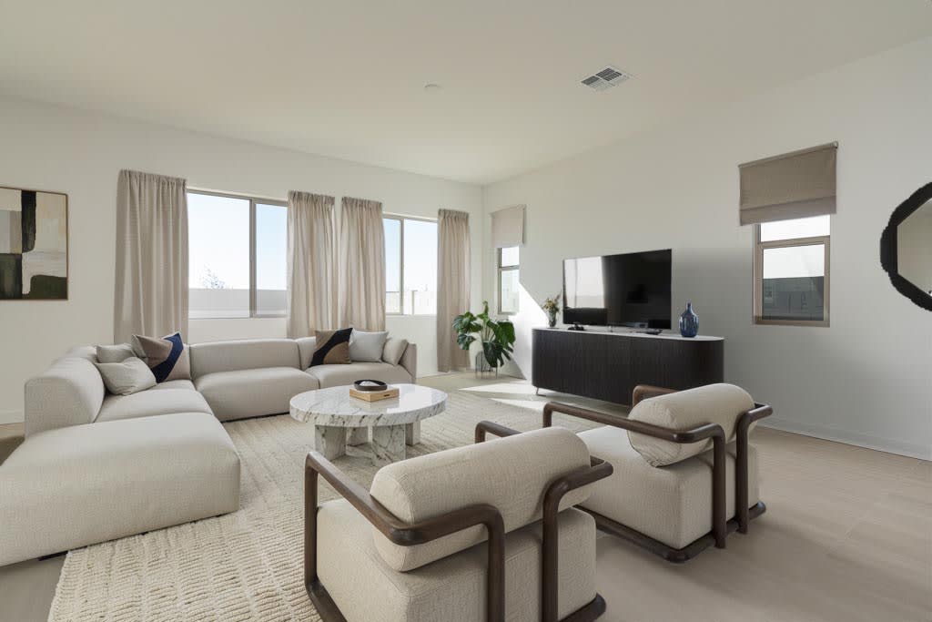 A modern living room features a white sectional sofa and two armchairs around a marble coffee table in the foreground, with a television console and large windows in the background.
