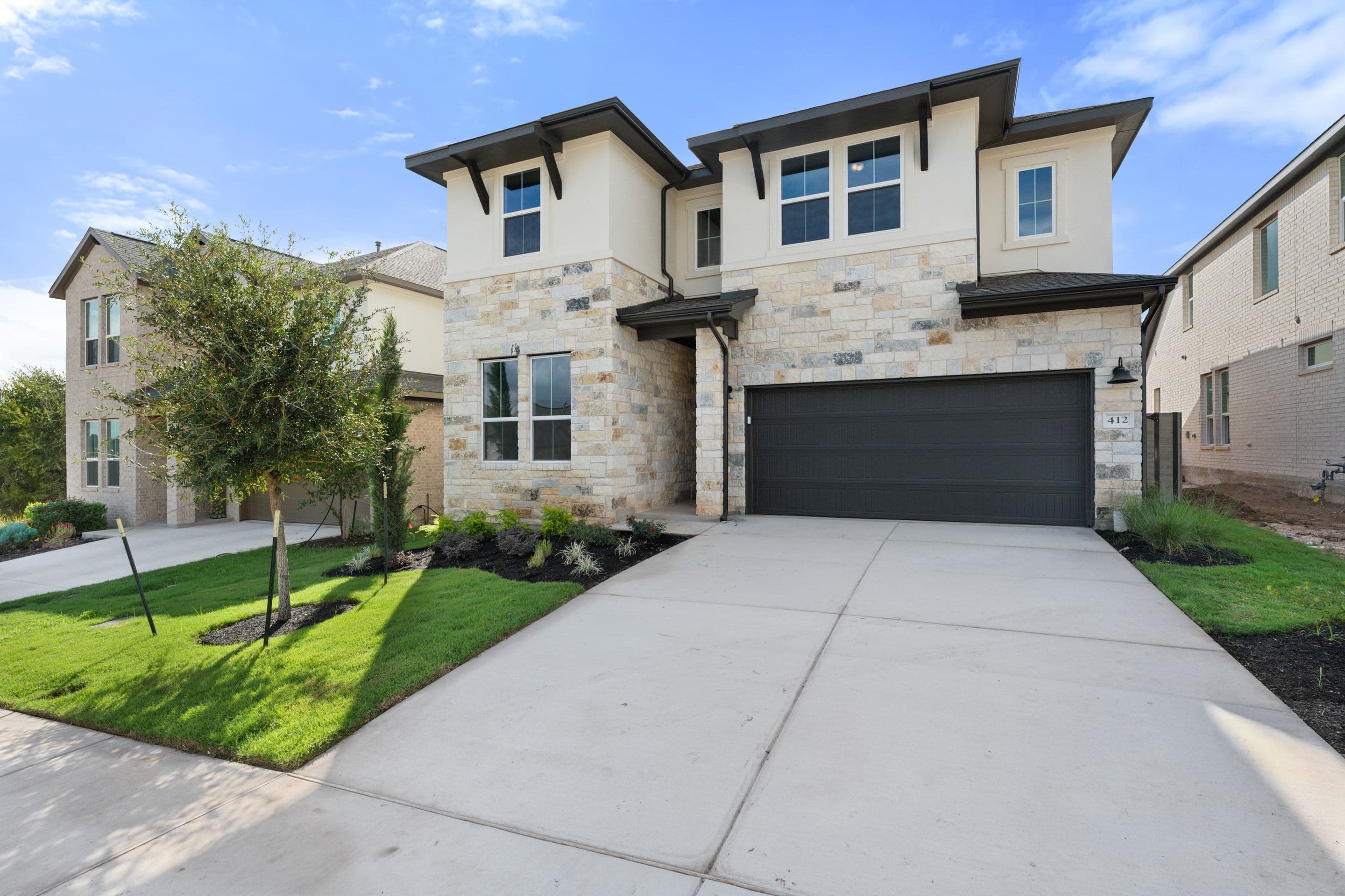 A modern two-story stone and stucco home with a dark garage door sits behind a concrete driveway and green lawn under a blue sky.