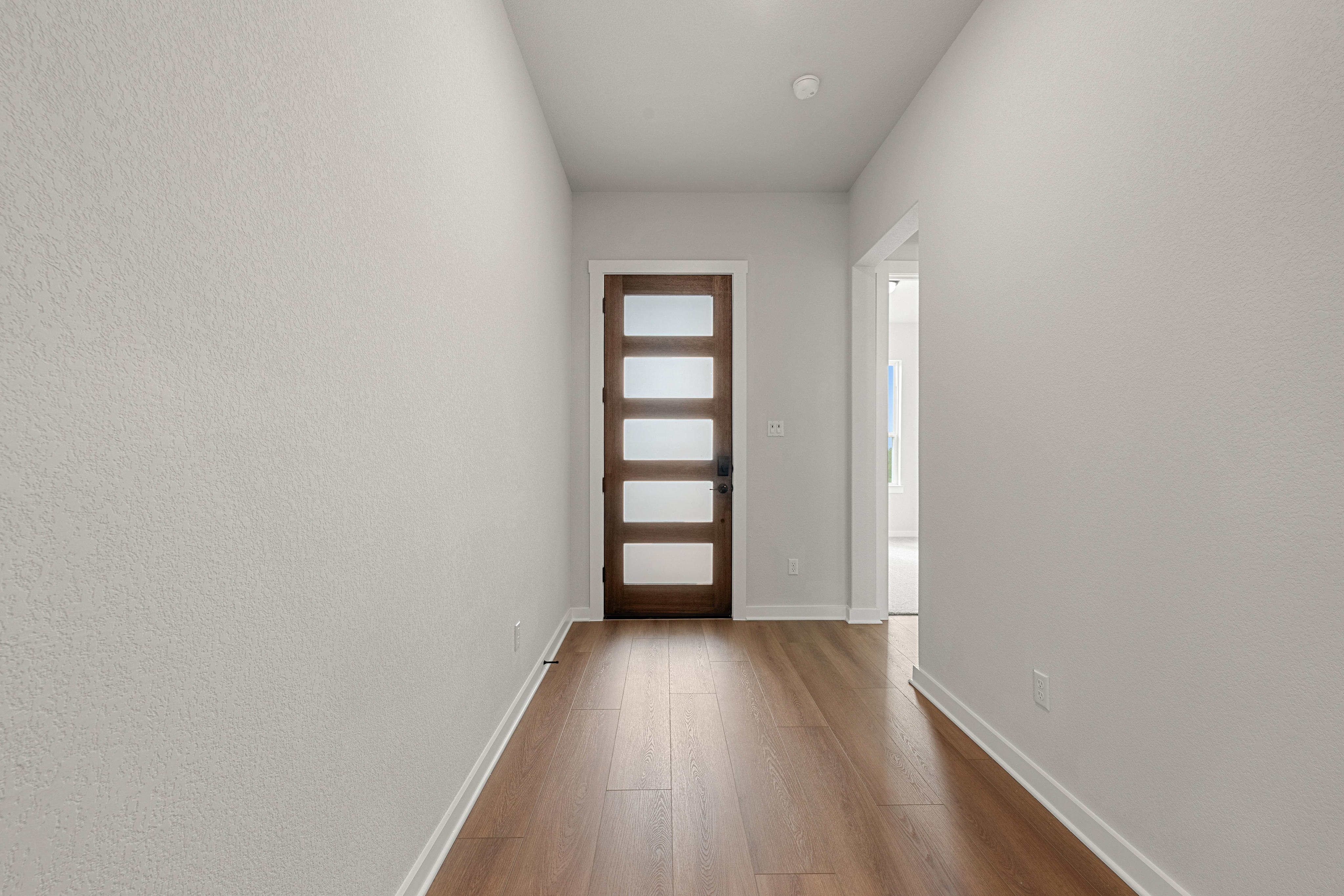 A narrow hallway with light wood flooring leads toward a dark wood door with frosted glass panels, flanked by plain white walls.