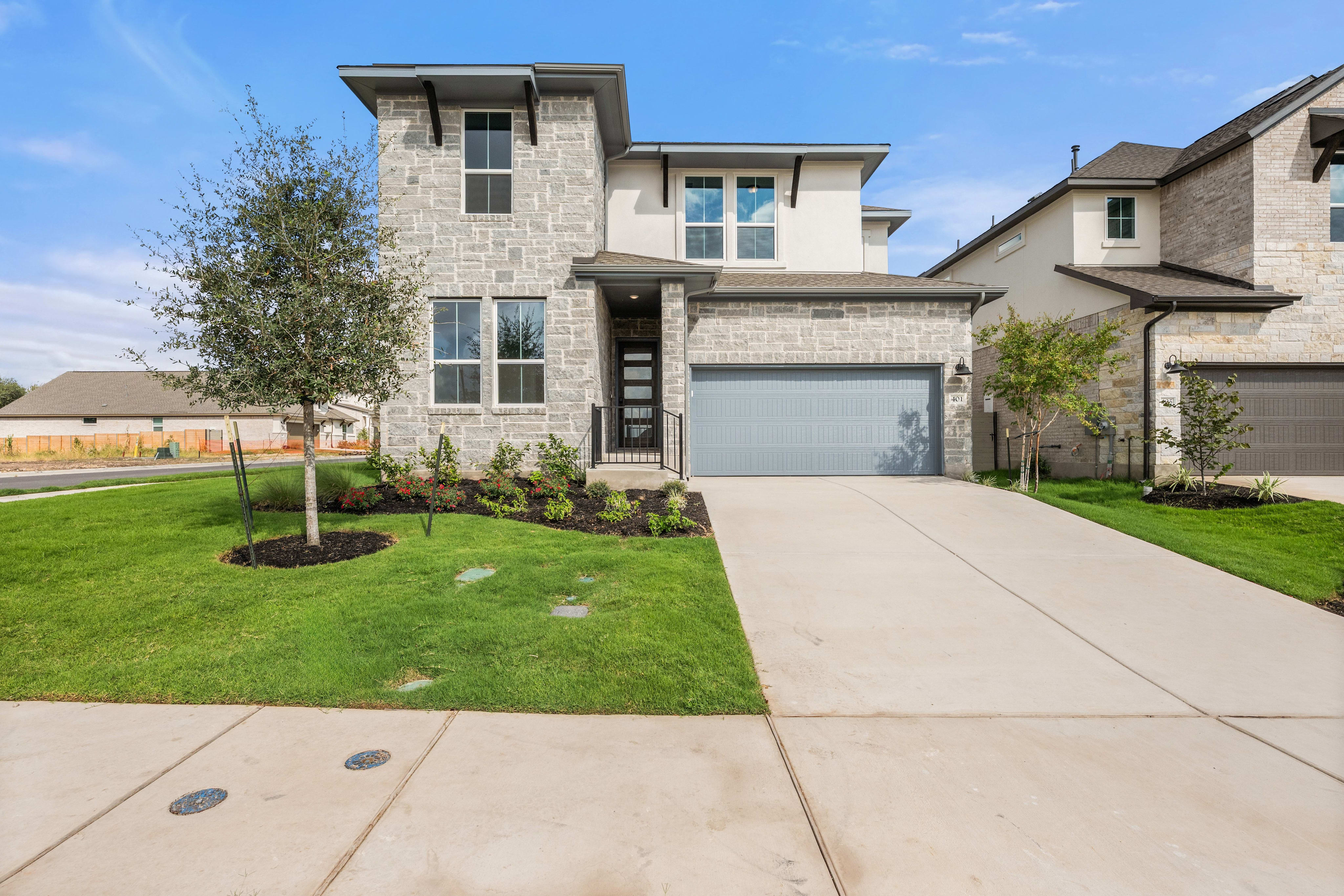 A modern stone house with a grey garage door and a green lawn in the foreground sits under a blue sky with a neighboring home in the background.