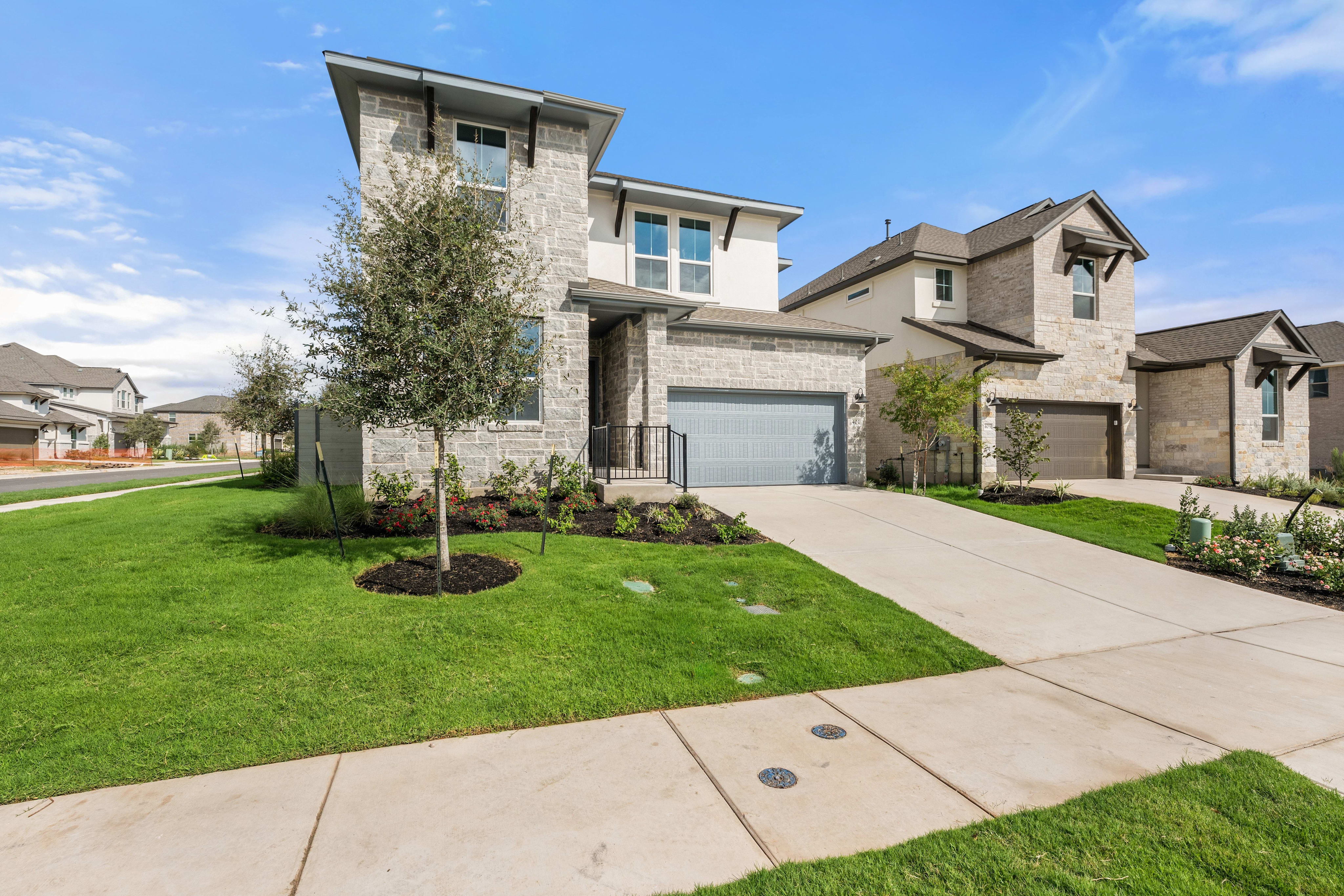 A modern stone house with a manicured lawn and concrete driveway sits under a bright blue sky with neighboring homes in the background.