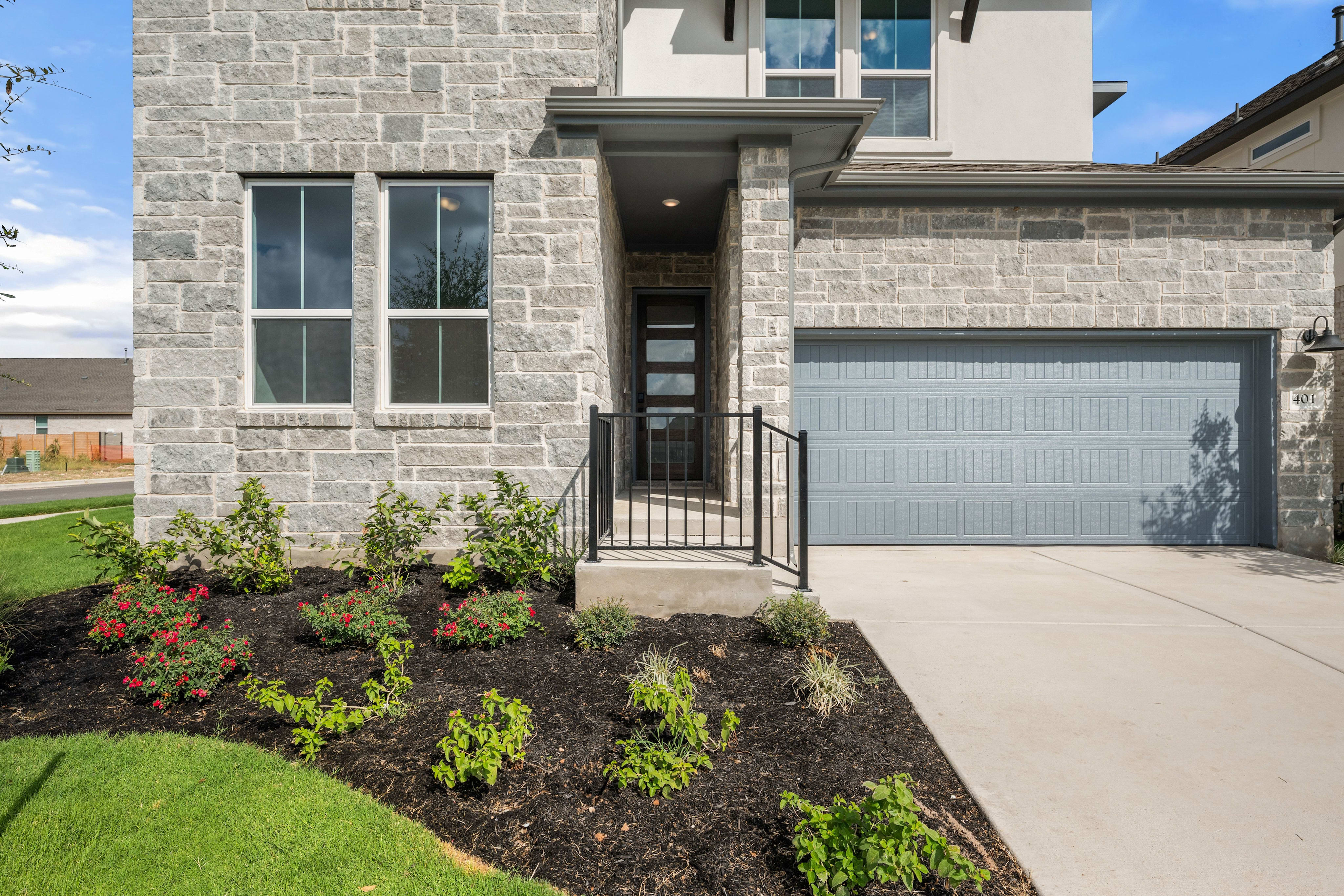 A grey stone house with a blue garage door stands behind a landscaped garden bed and a concrete driveway.