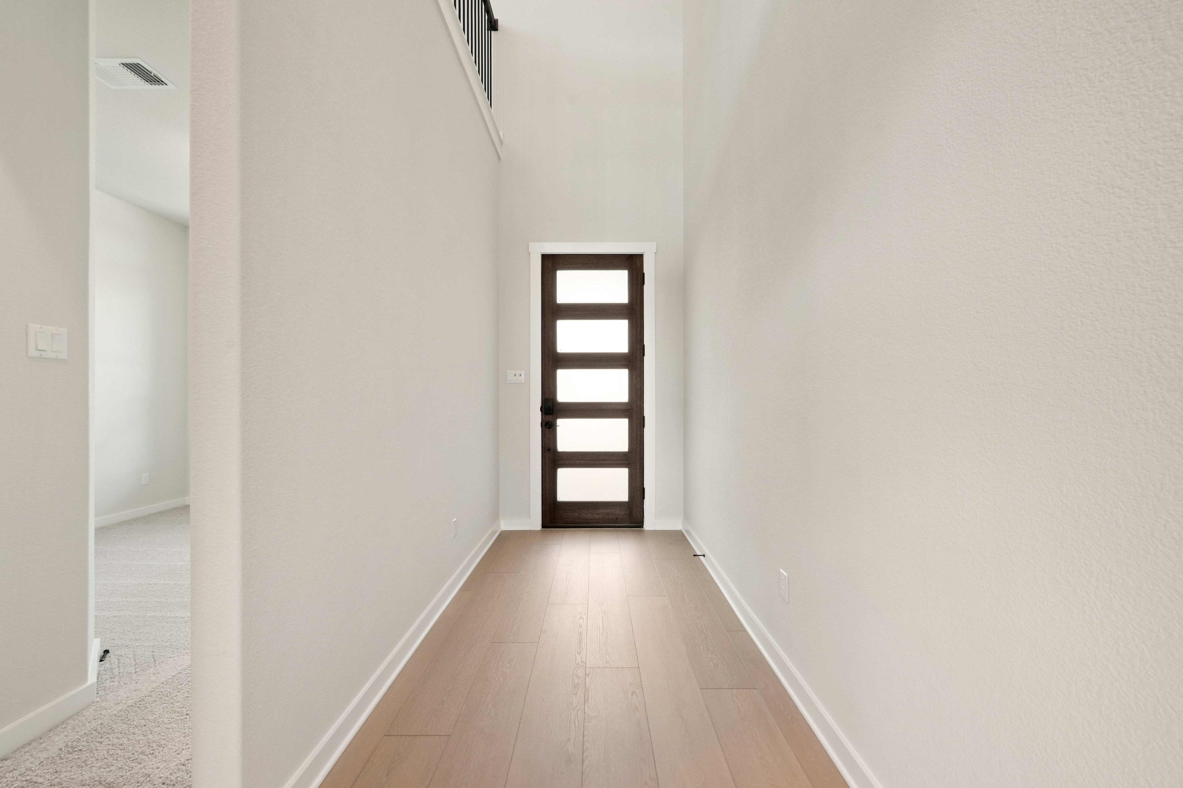 A light-colored wooden hallway leads toward a dark brown door with horizontal glass panes, flanked by white walls.