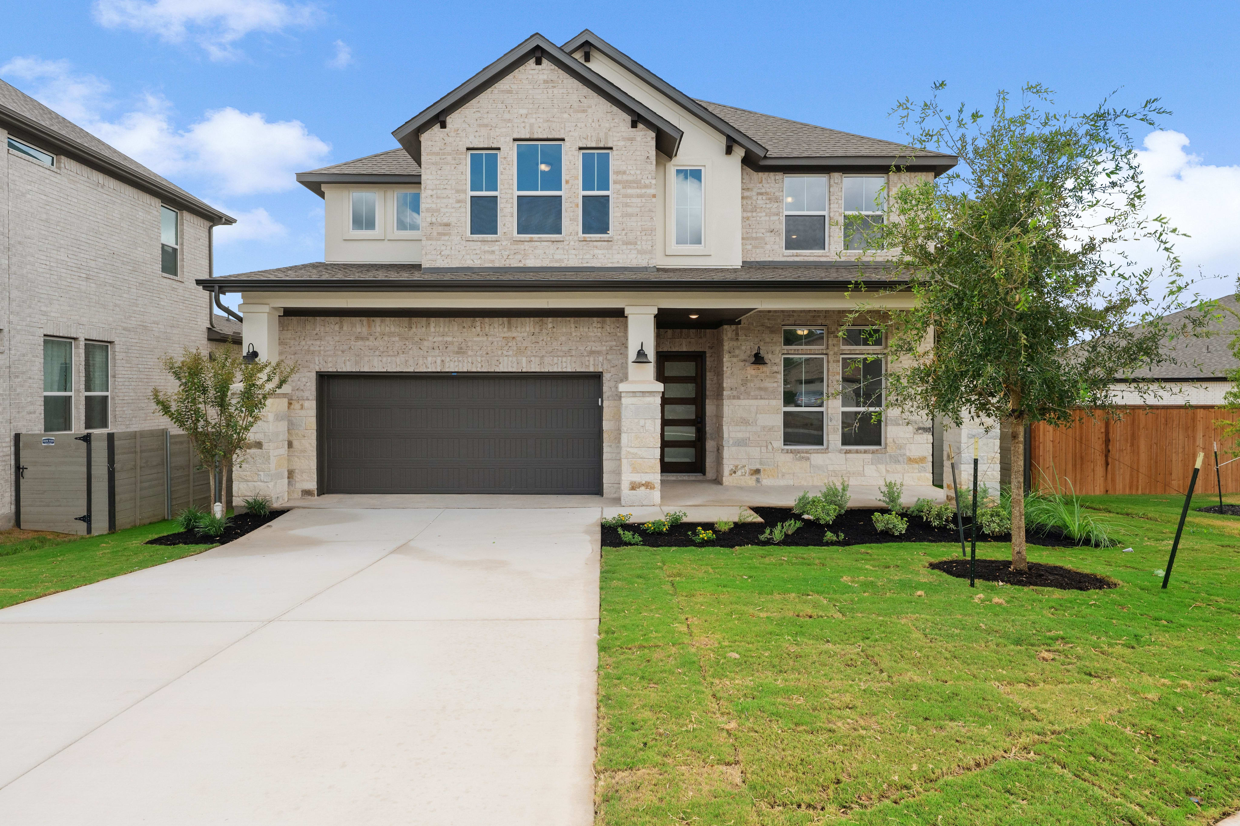A modern two-story stone house with a grey garage door stands behind a concrete driveway and green lawn under a blue sky.