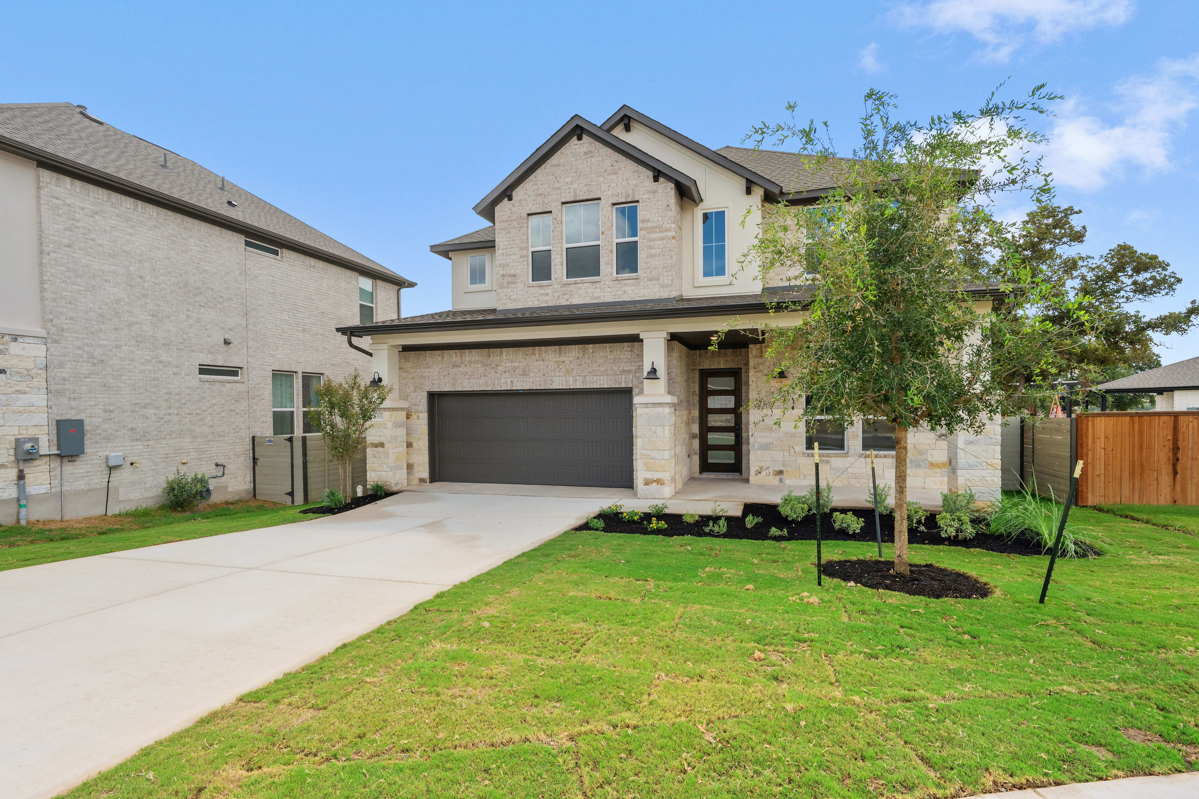 A concrete driveway and green lawn lead to a modern two-story stone house under a bright blue sky.