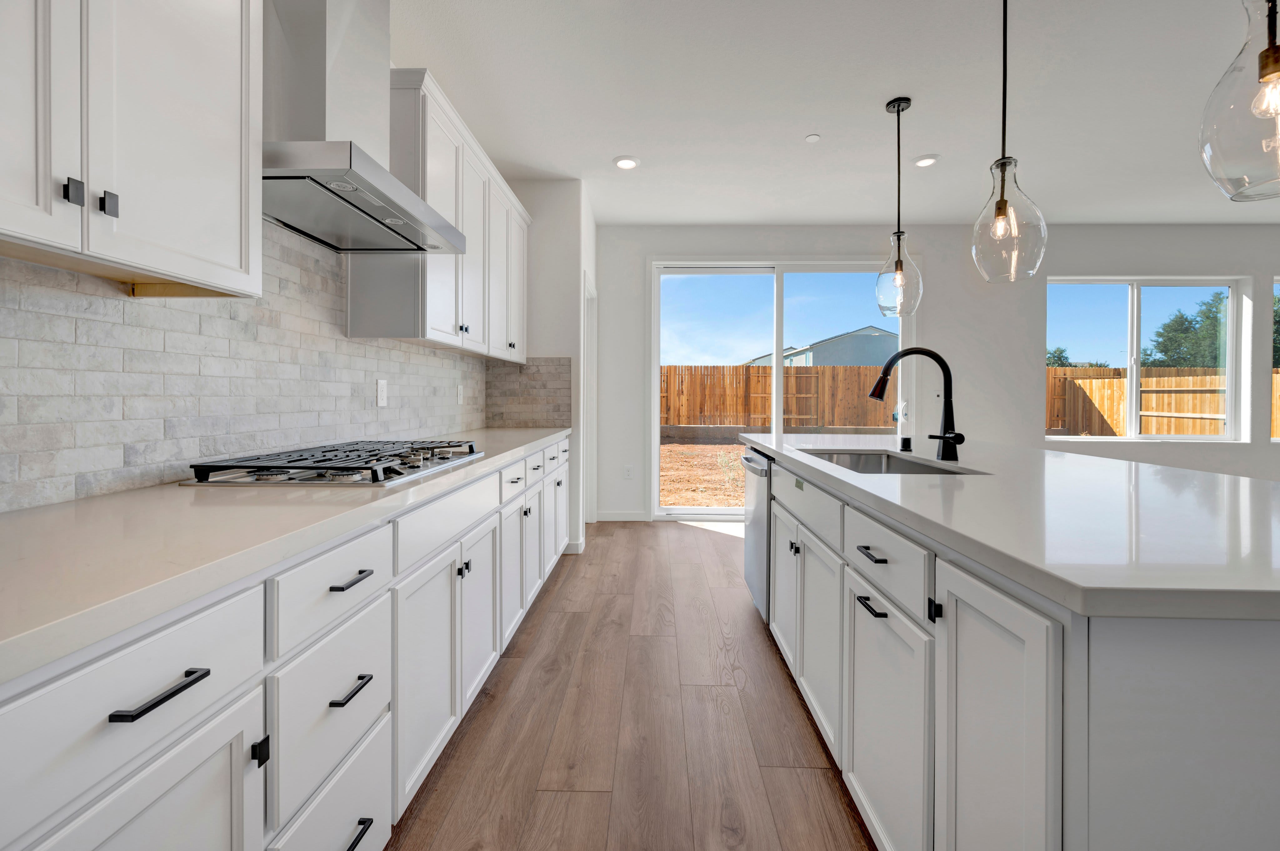 A modern white kitchen with a large island and gas stovetop opens up to a bright backyard view through a sliding glass door.