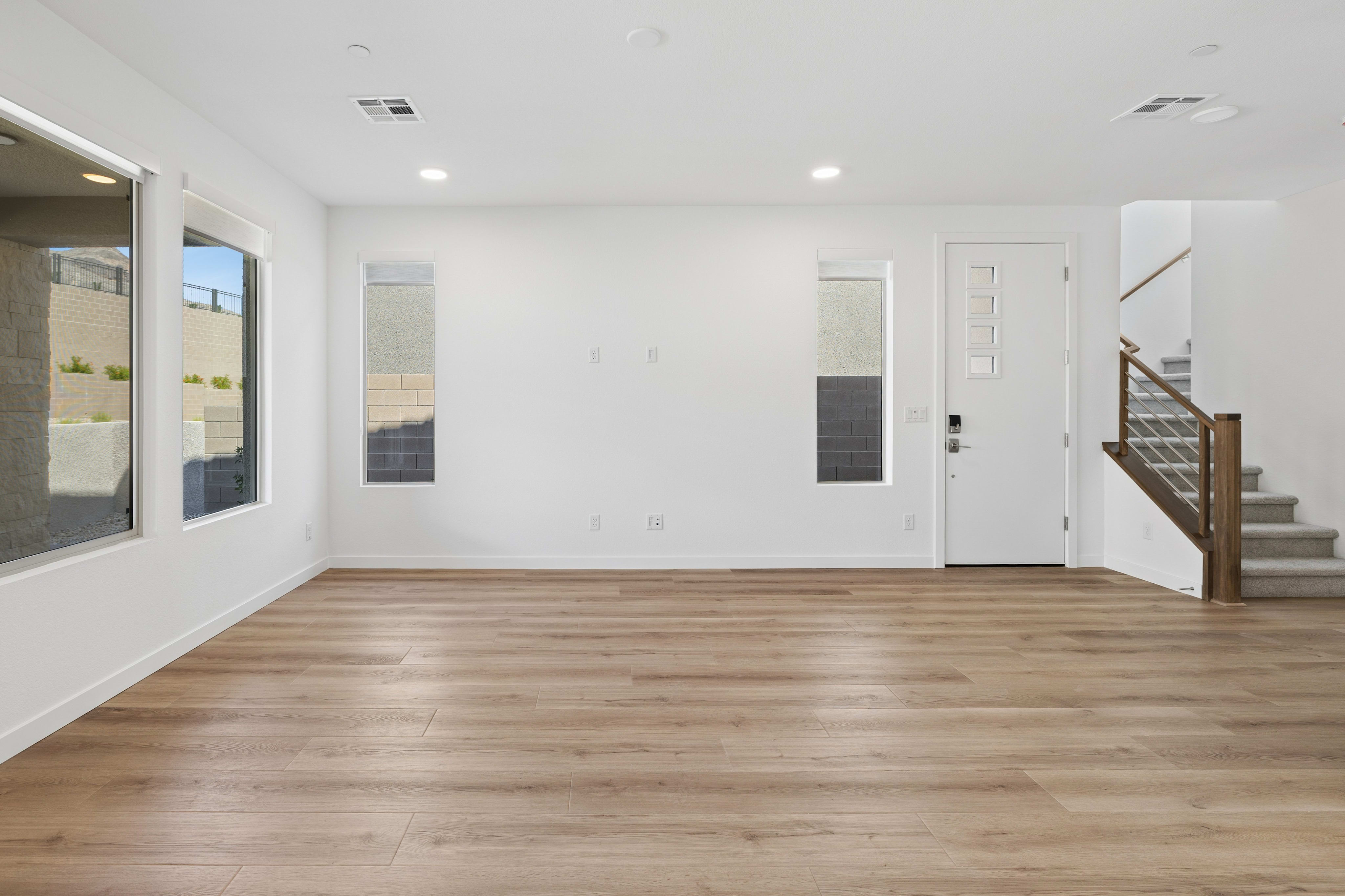 An empty room with light wood flooring in the foreground and white walls with windows and a door leading to a staircase in the background.