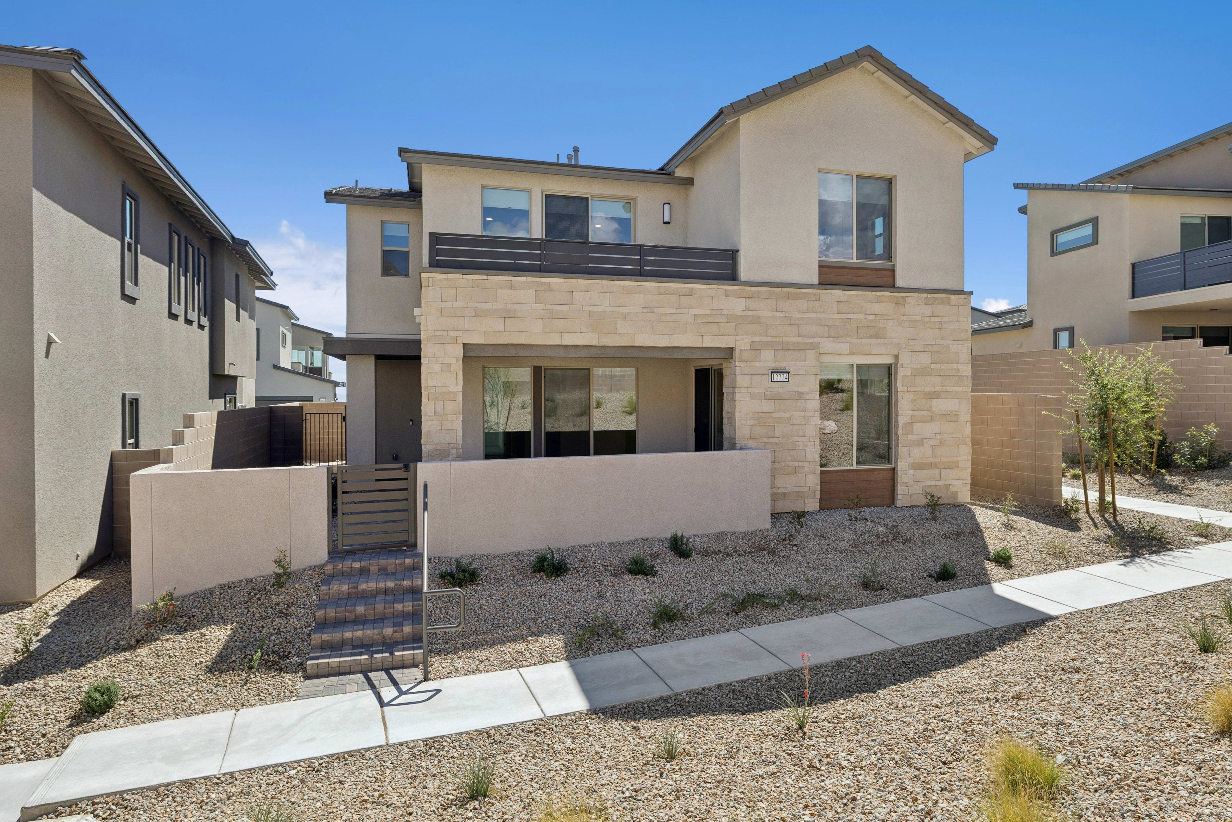 A modern two-story home with a stone facade and balcony stands behind a gravel yard and concrete walkway under a clear blue sky.