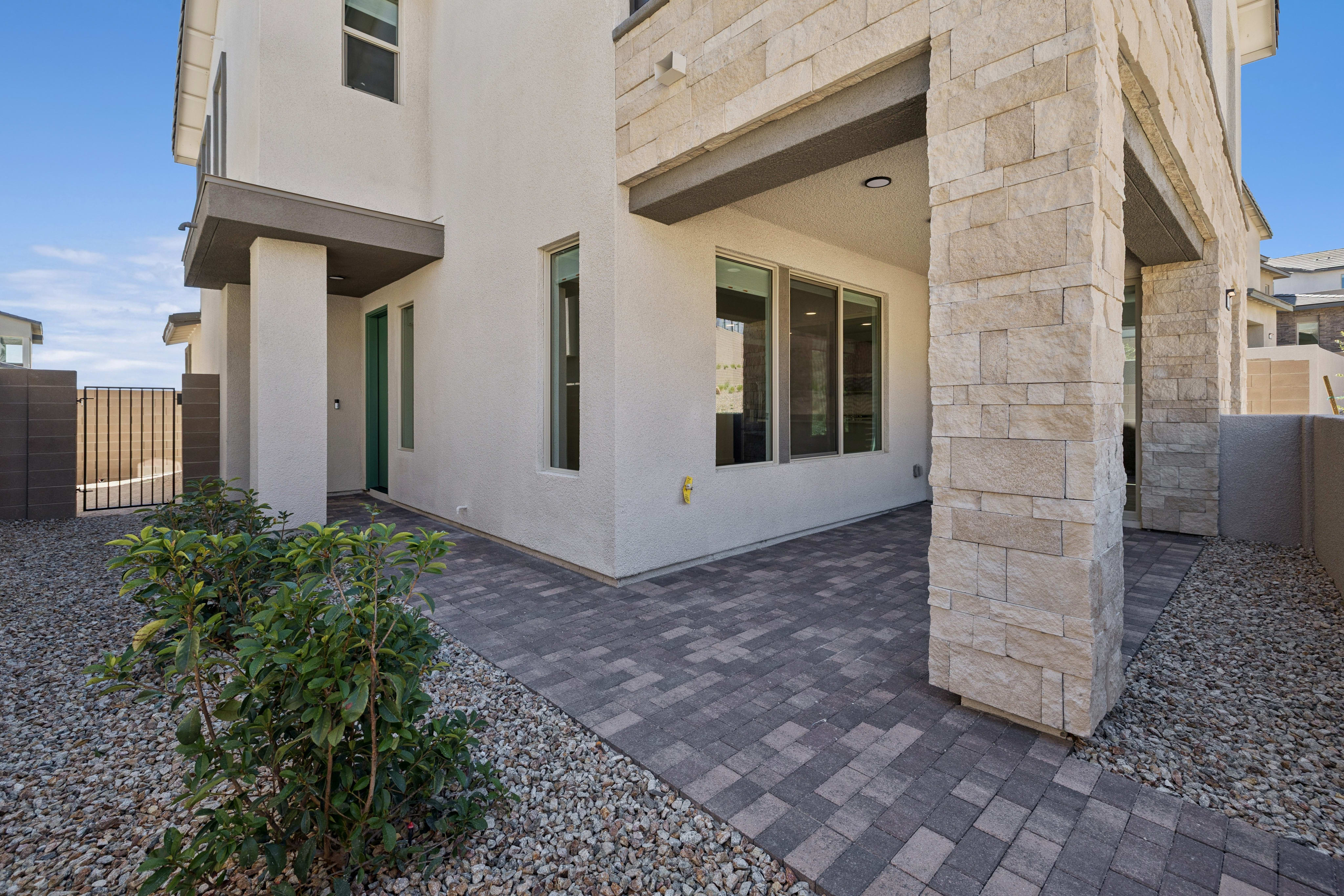 A green shrub sits in a gravel yard in the foreground, leading to a paved patio and a modern house with stone accents under a clear blue sky.