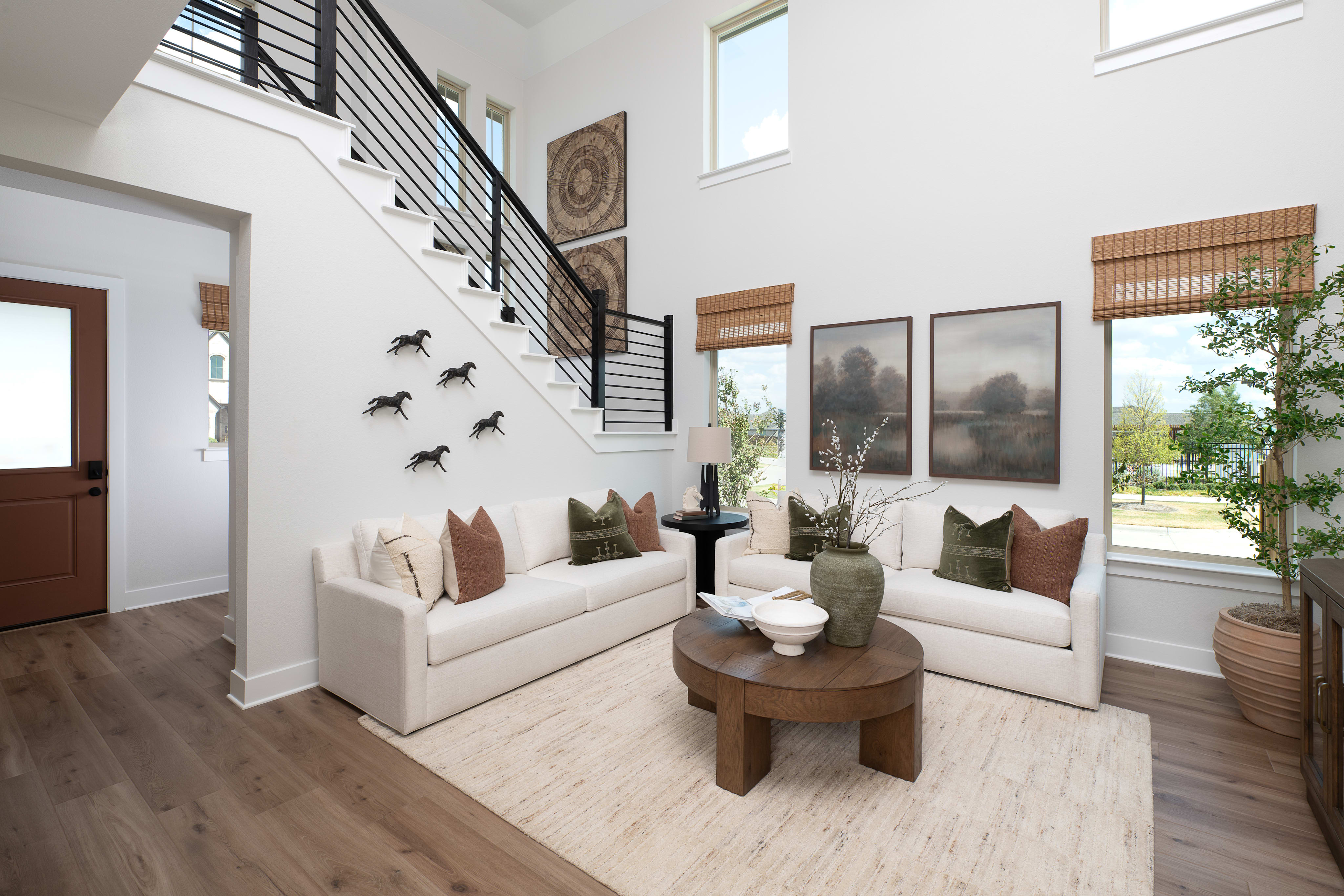 A modern living room features white sofas and a round wooden coffee table in the foreground, with a staircase and large windows in the background.
