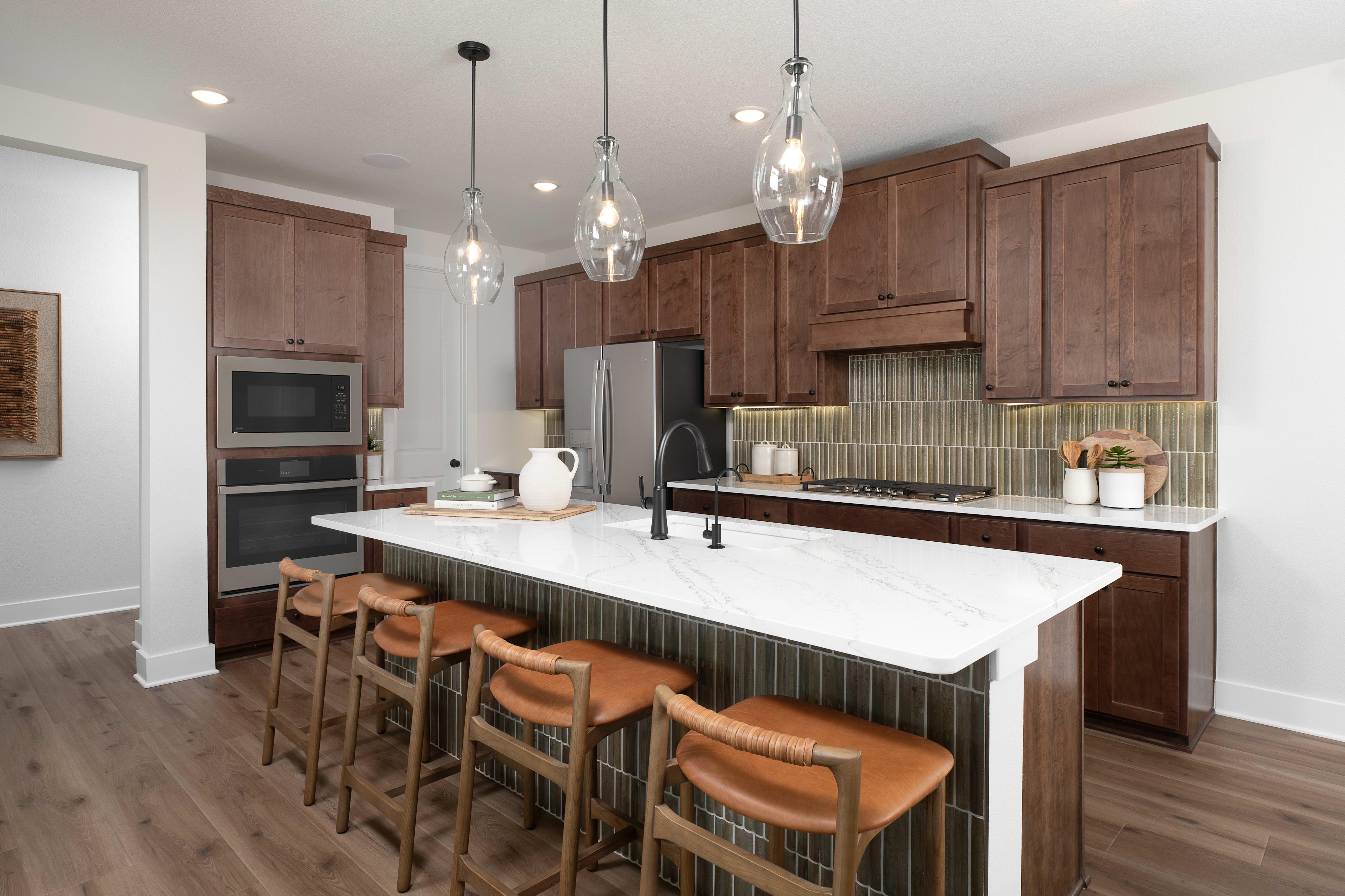 A white marble kitchen island with leather-seated barstools sits in the foreground of a modern kitchen featuring dark wood cabinetry and a green tiled backsplash.