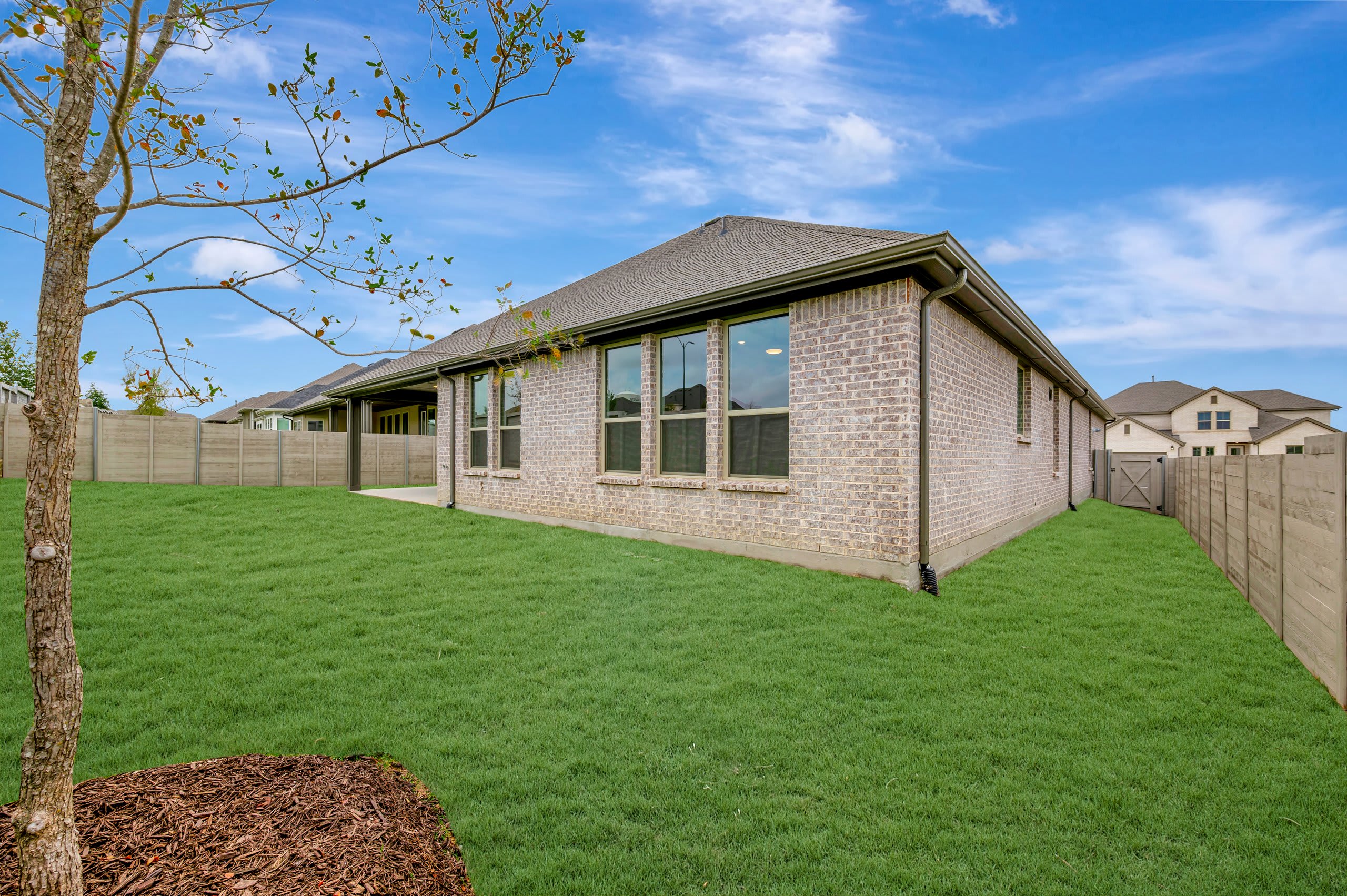 A young tree and green lawn occupy the foreground, leading to a light-colored brick house set against a bright blue sky with wispy clouds.