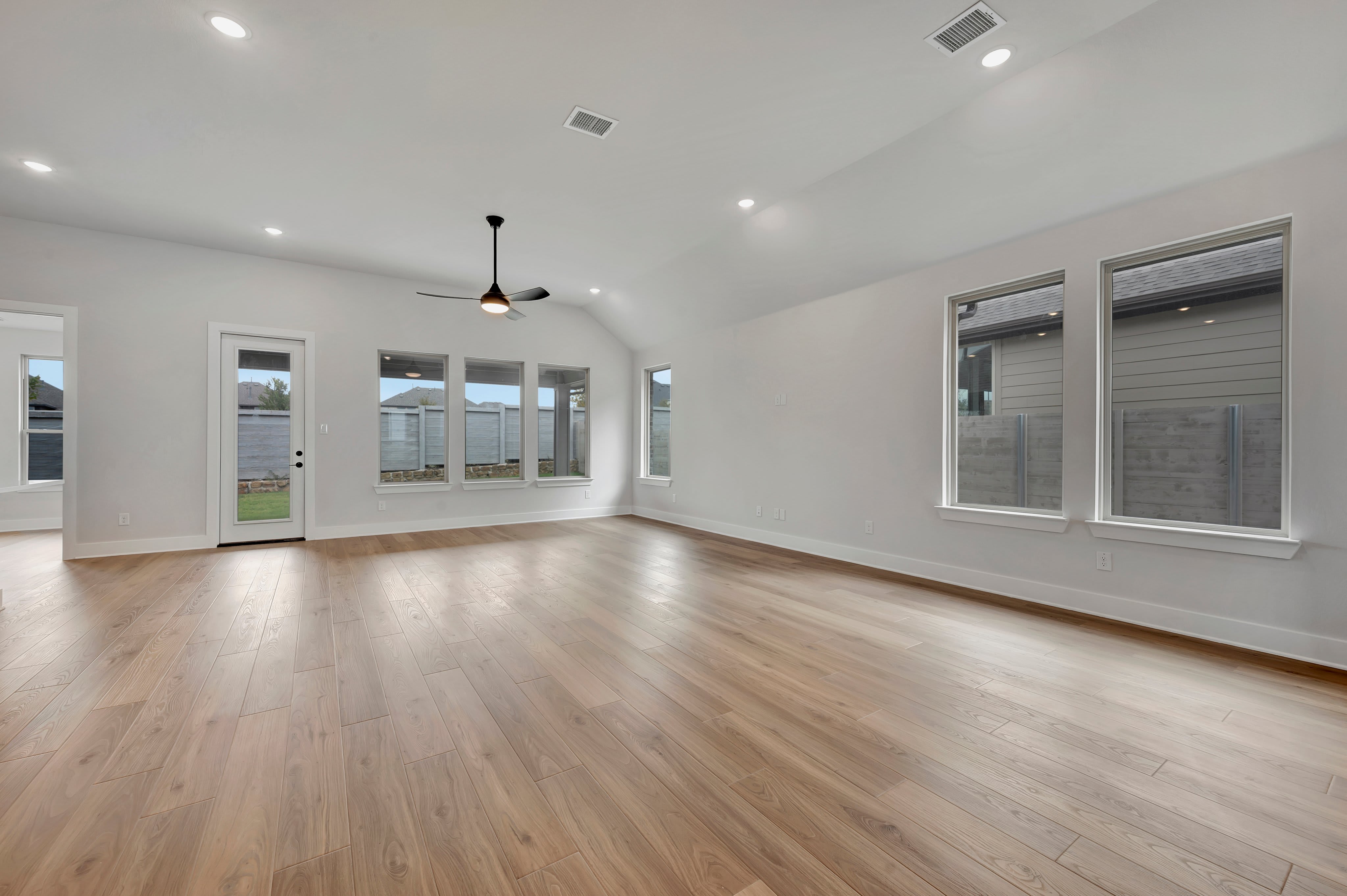 An empty room with light wood flooring in the foreground and white walls with several windows and a ceiling fan in the background.