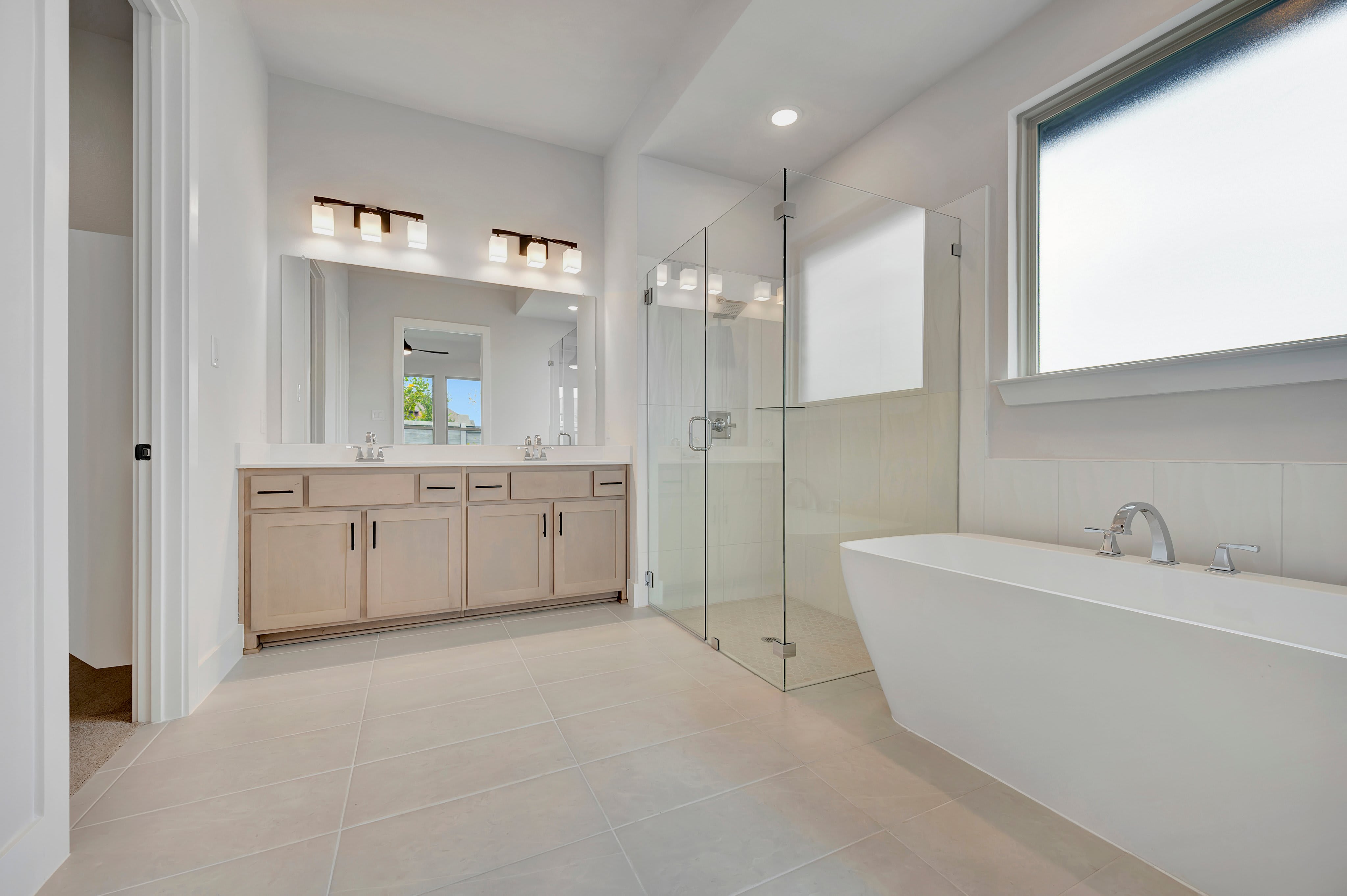 A modern white bathtub sits in the foreground of a bright bathroom featuring a glass shower and a double vanity in the background.