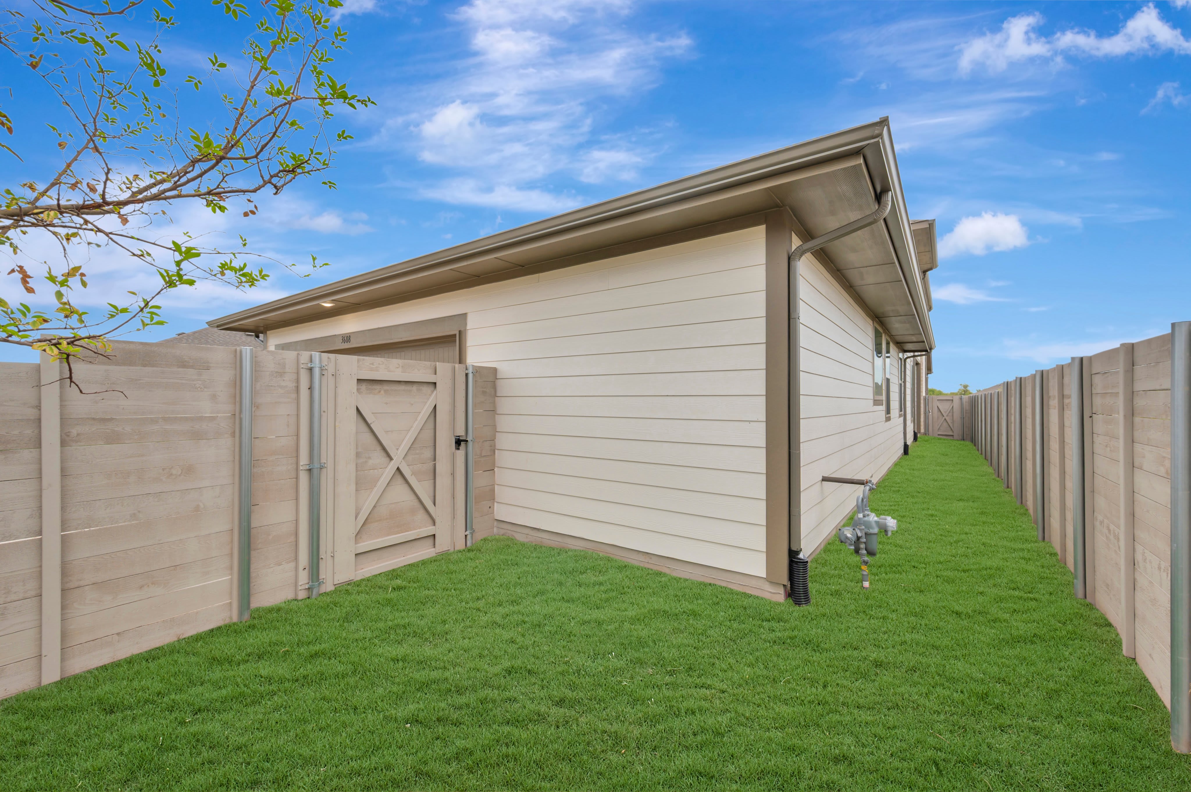 A manicured green lawn is enclosed by a light-colored wooden fence and the side of a modern house under a bright blue sky with wispy clouds.