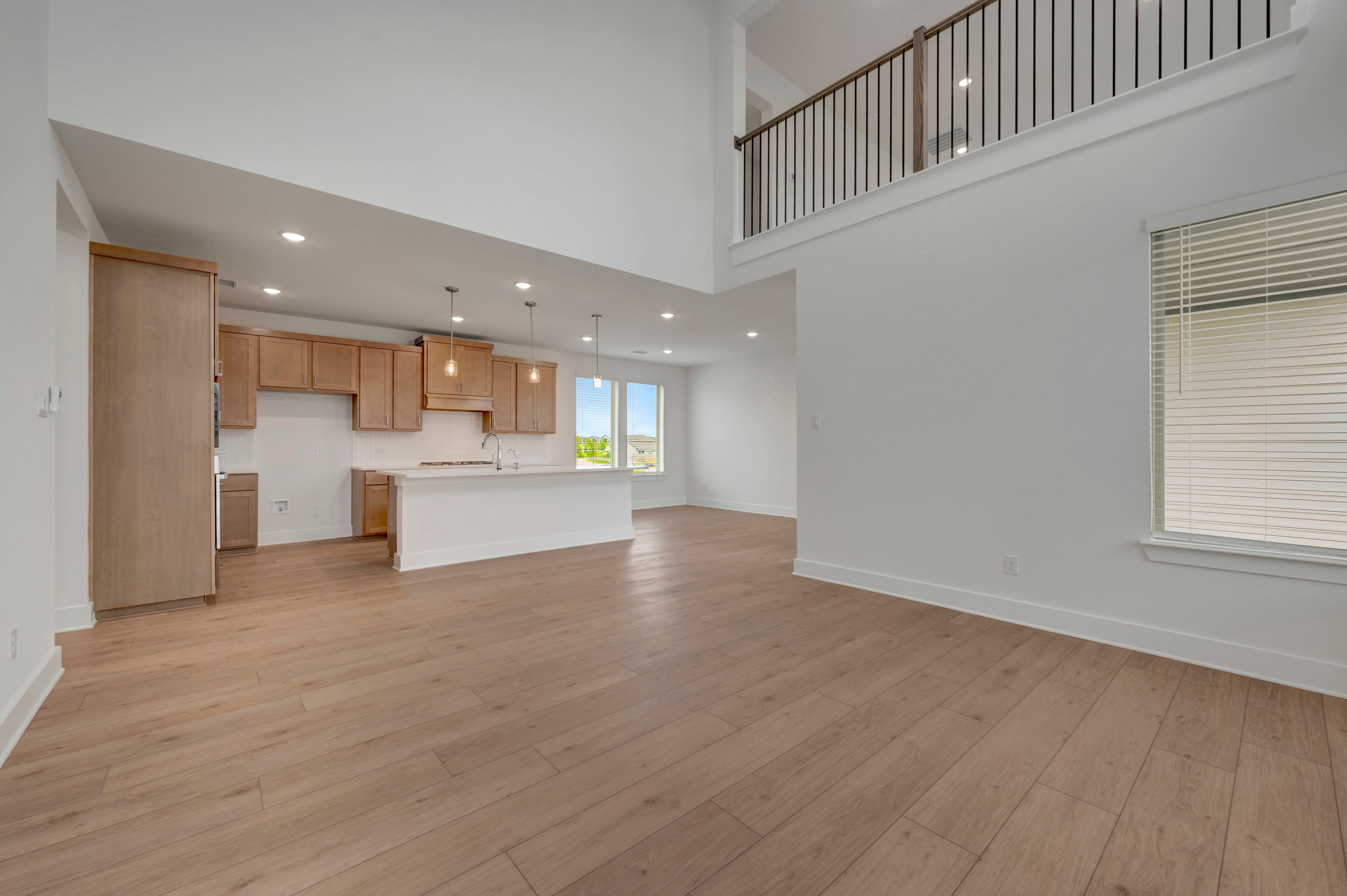 Light-colored wood flooring fills the foreground of an open-concept living area, leading back to a kitchen with wooden cabinets and a white island beneath a second-floor balcony.
